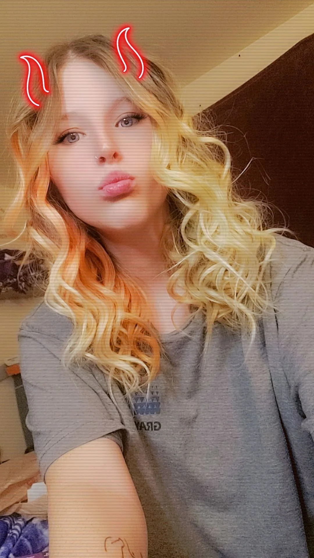A woman with long, curly blonde hair, making a pouty face with her lips. She has digital red devil horns and whiskers added to her face. She is wearing a gray shirt and a necklace, in an indoor setting.