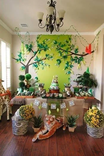Lion plush toy sitting in front of a decorated table for a child's birthday party with a jungle theme, featuring cake, balloons, plants, and jungle animal decorations on a green wall with a branch and leaf mural.