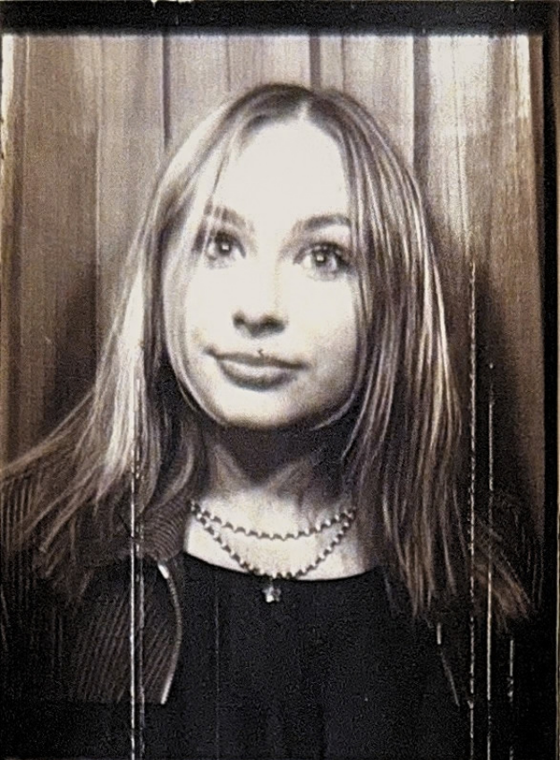 Black and white photo of a young woman with long hair, wearing a necklace, and looking slightly to the side, with a wooden background.
