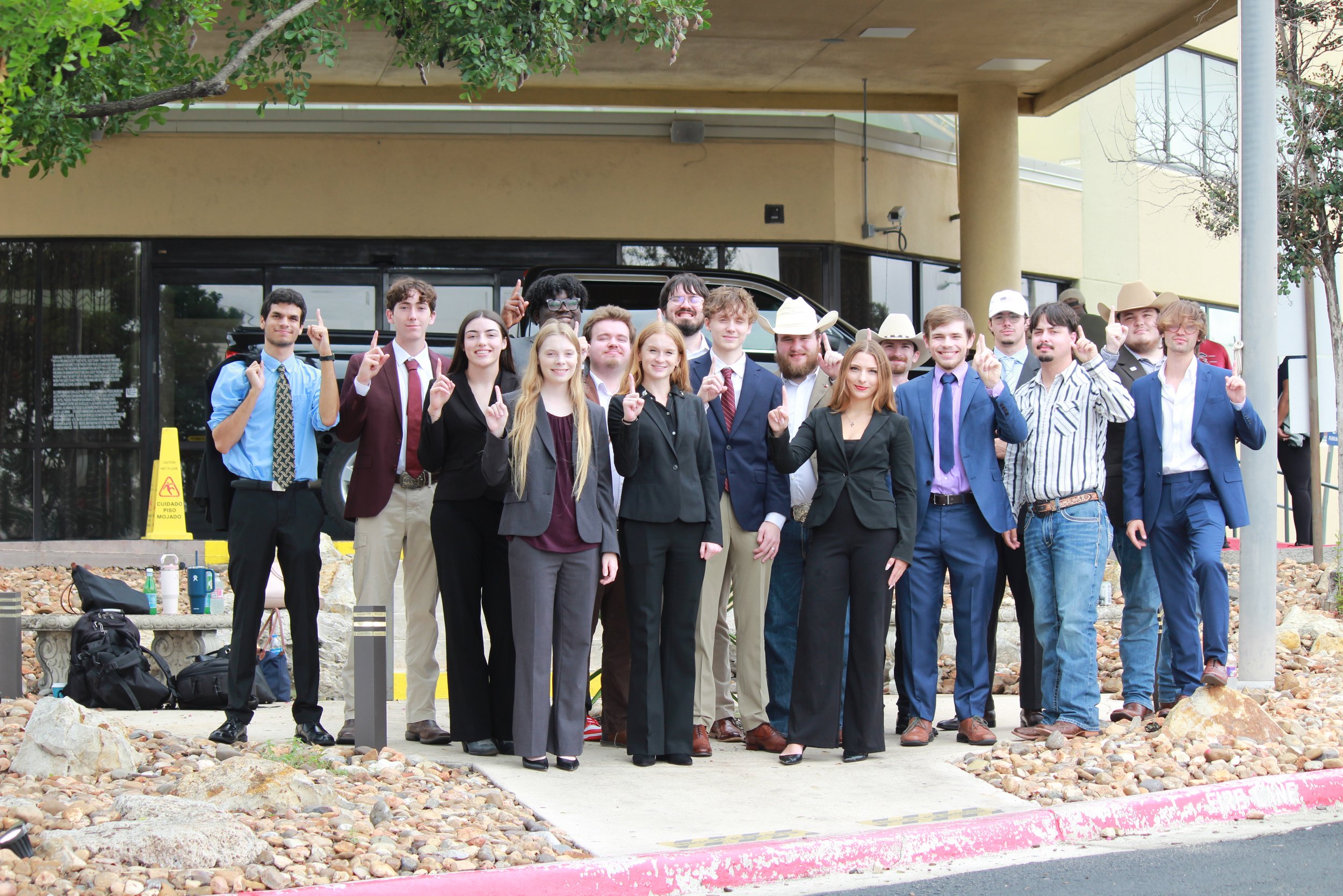 Group of young adults, dressed in semi-formal attire, standing outside a building, smiling and raising their index fingers. They are standing on a sidewalk with rocks, bags, and water bottles in the foreground. The background shows a beige building with large windows and some trees.