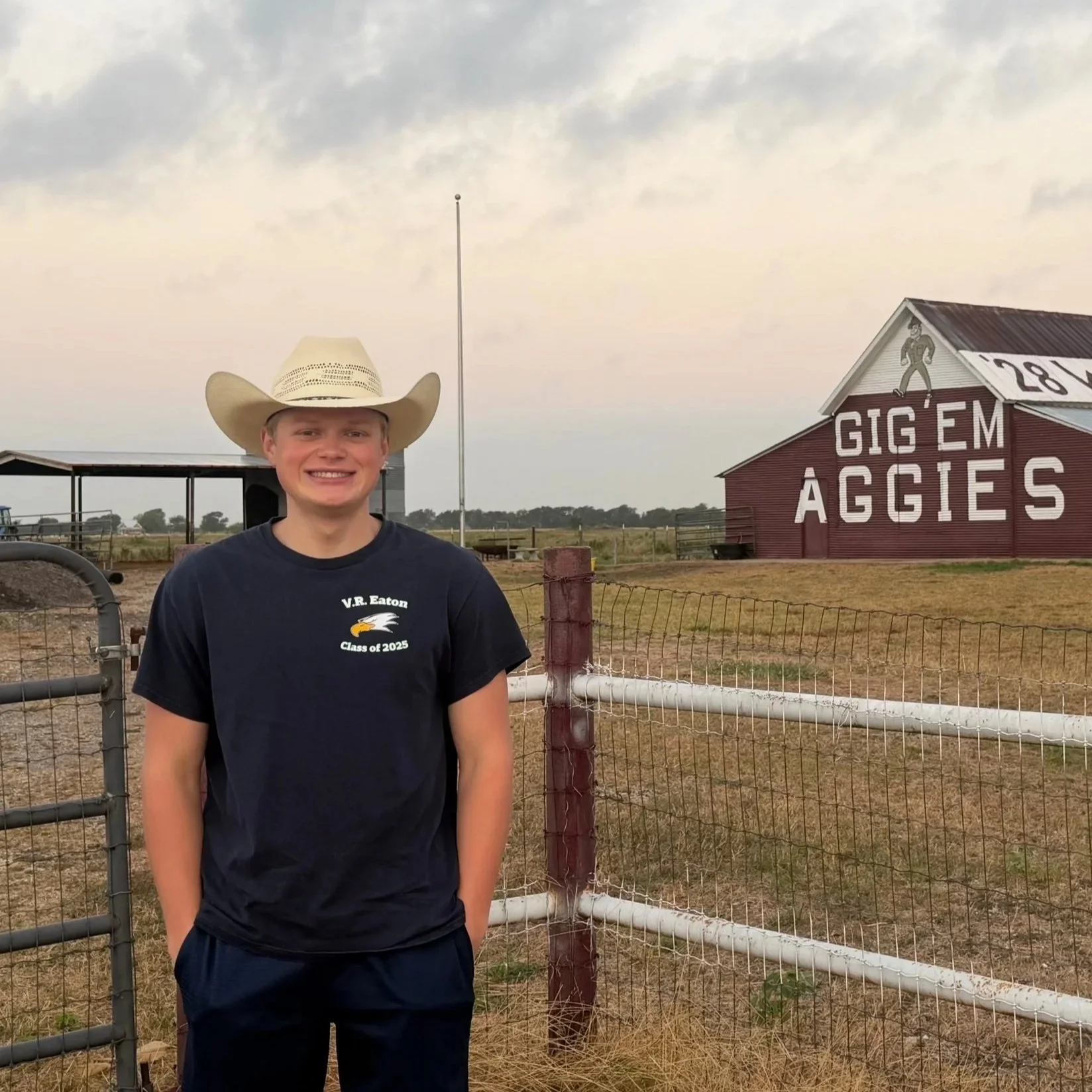 Young man wearing a cowboy hat and a black t-shirt with a graphic and text stands outdoors in front of a rural farm with a red barn that has large white letters reading "GIG EM AGGIES" and an illustration of a cowboy, under a cloudy sky.