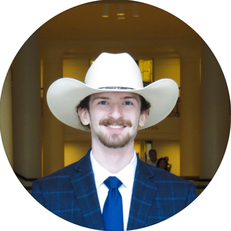 A man with a beard and mustache wearing a white cowboy hat, blue suit, and blue tie, smiling at the camera in an indoor setting.