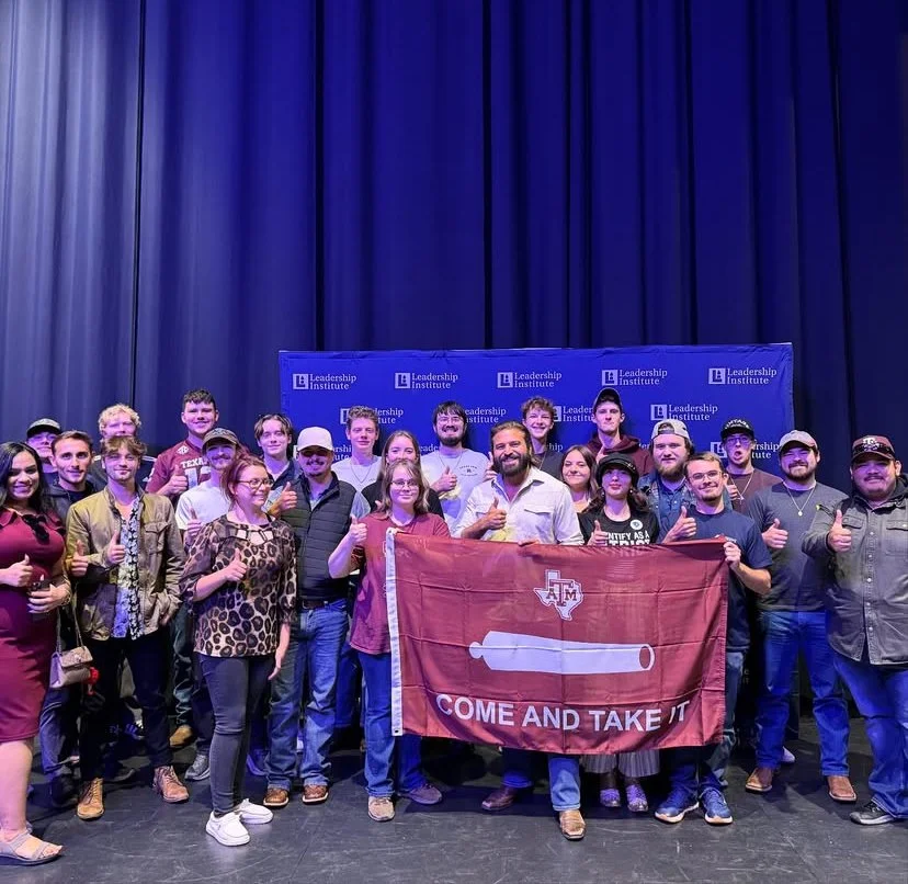 Group of people on stage at the Leadership Institute, holding a red flag with a white cannon and Texas outline, and the text 'Come and Take It'.