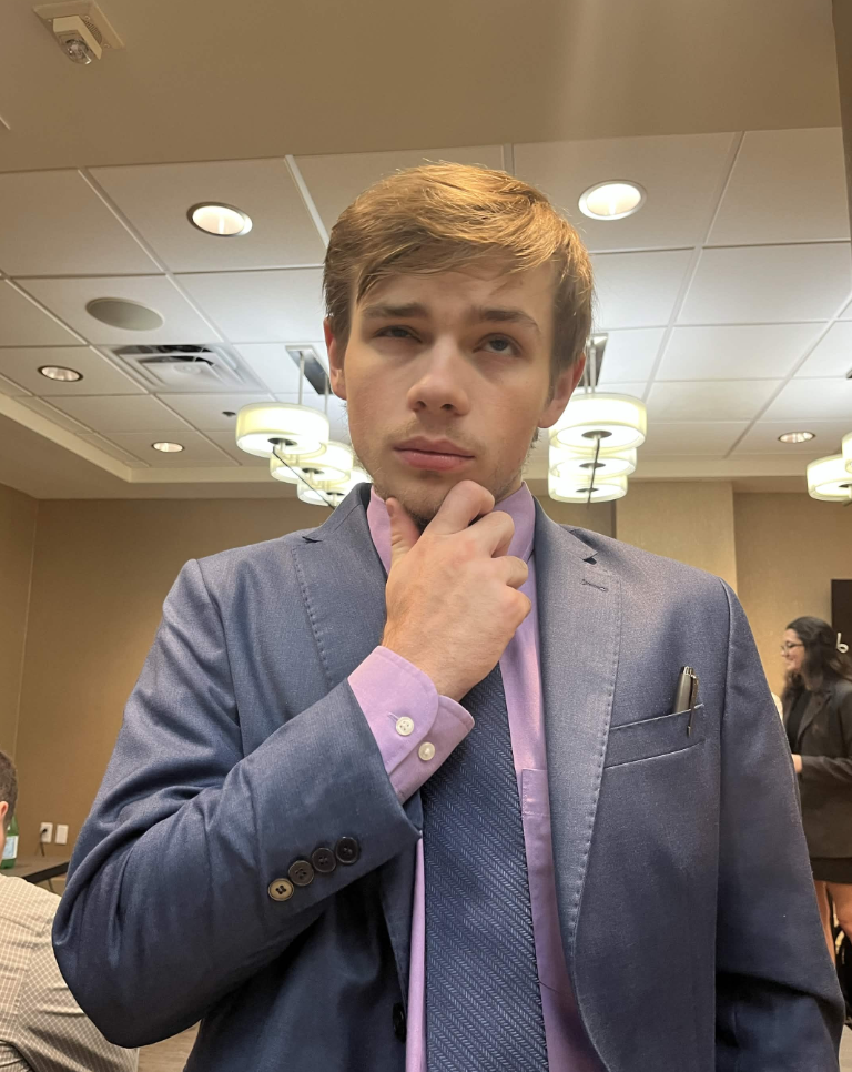 A young man in a blue suit and purple shirt, pondering with his hand on his chin in a conference room or hotel lounge setting, with other people and ceiling lights visible in the background.