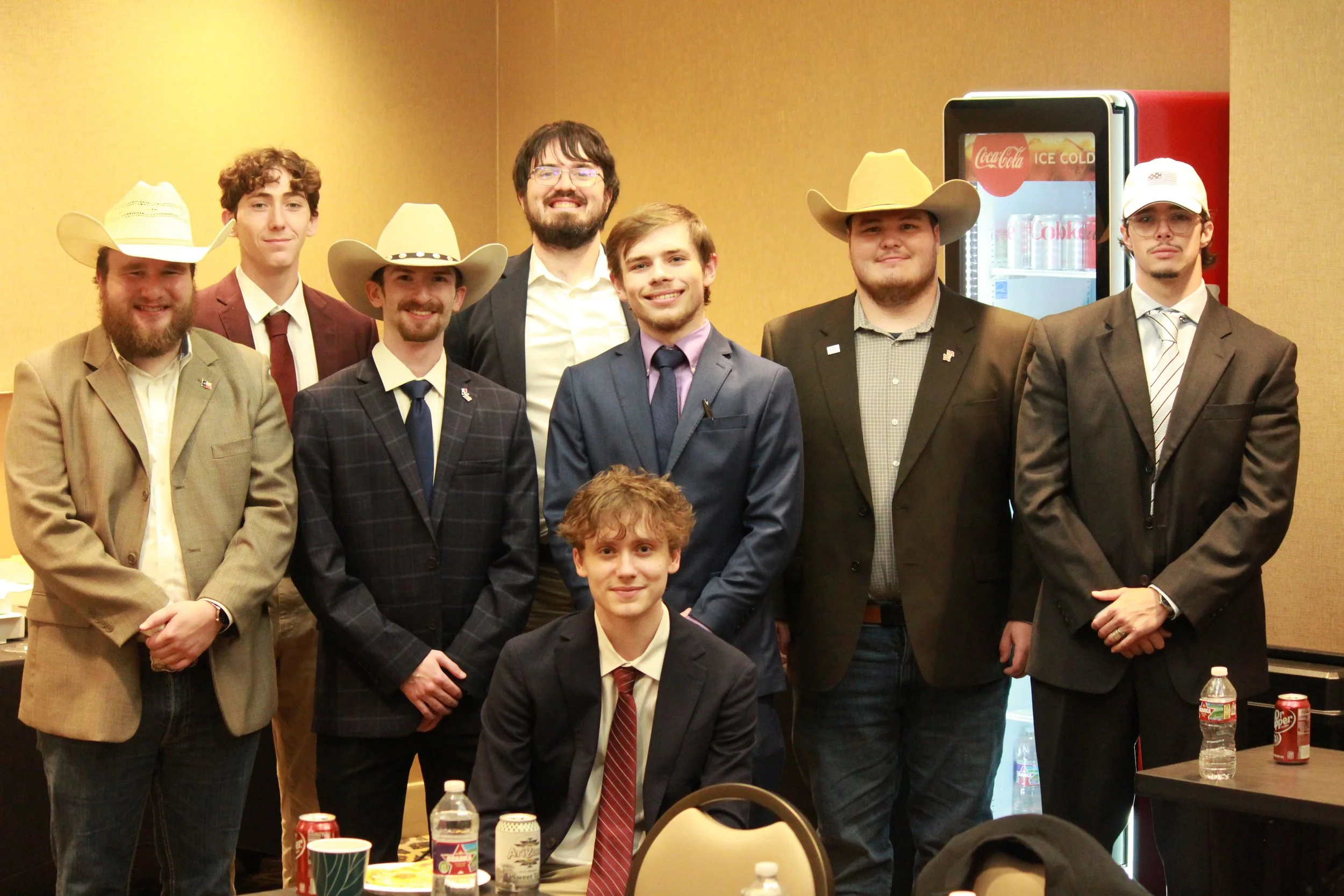 Group of seven men dressed in suits, some wearing cowboy hats, posing for a photo at a conference or event in a hotel or conference room with a beige wall and a Coca-Cola vending machine in the background. A table in front has drinks and snacks.
