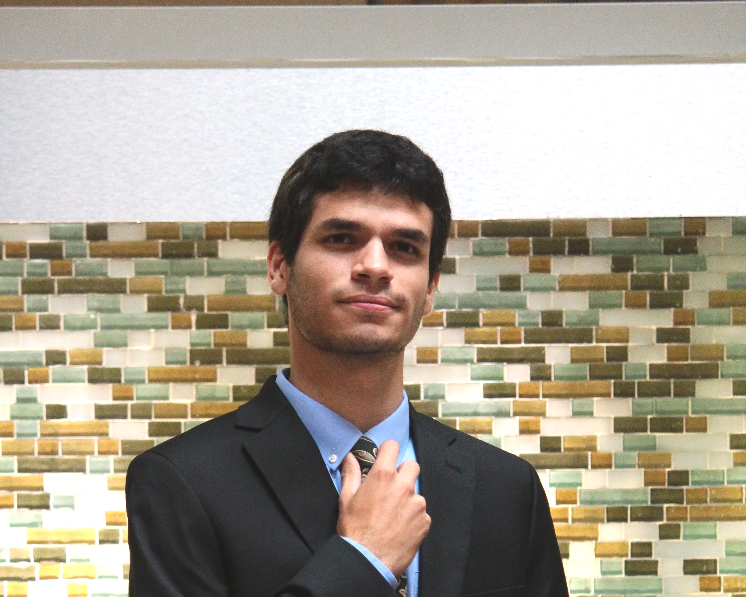 A young man in a suit with a tie, standing in front of a colorful mosaic wall, adjusting his tie with his left hand.