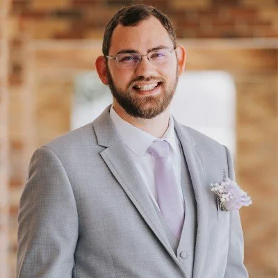 A man with glasses, a beard, and short dark hair smiling, wearing a light gray suit with a lavender tie and a boutonniere, in an indoor setting with wooden decor.