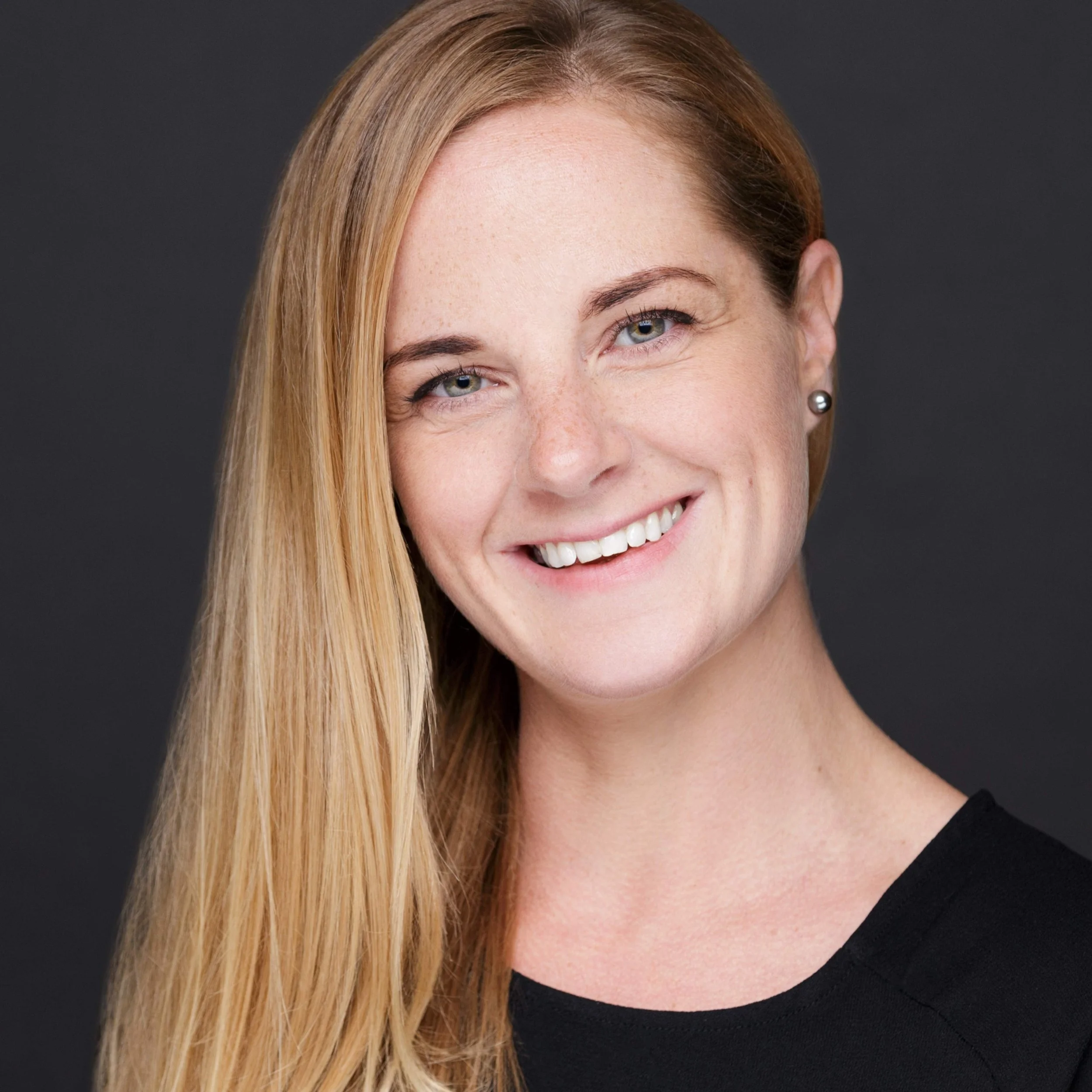 Close-up of a woman with long blonde hair, fair skin, and blue eyes smiling against a dark background.