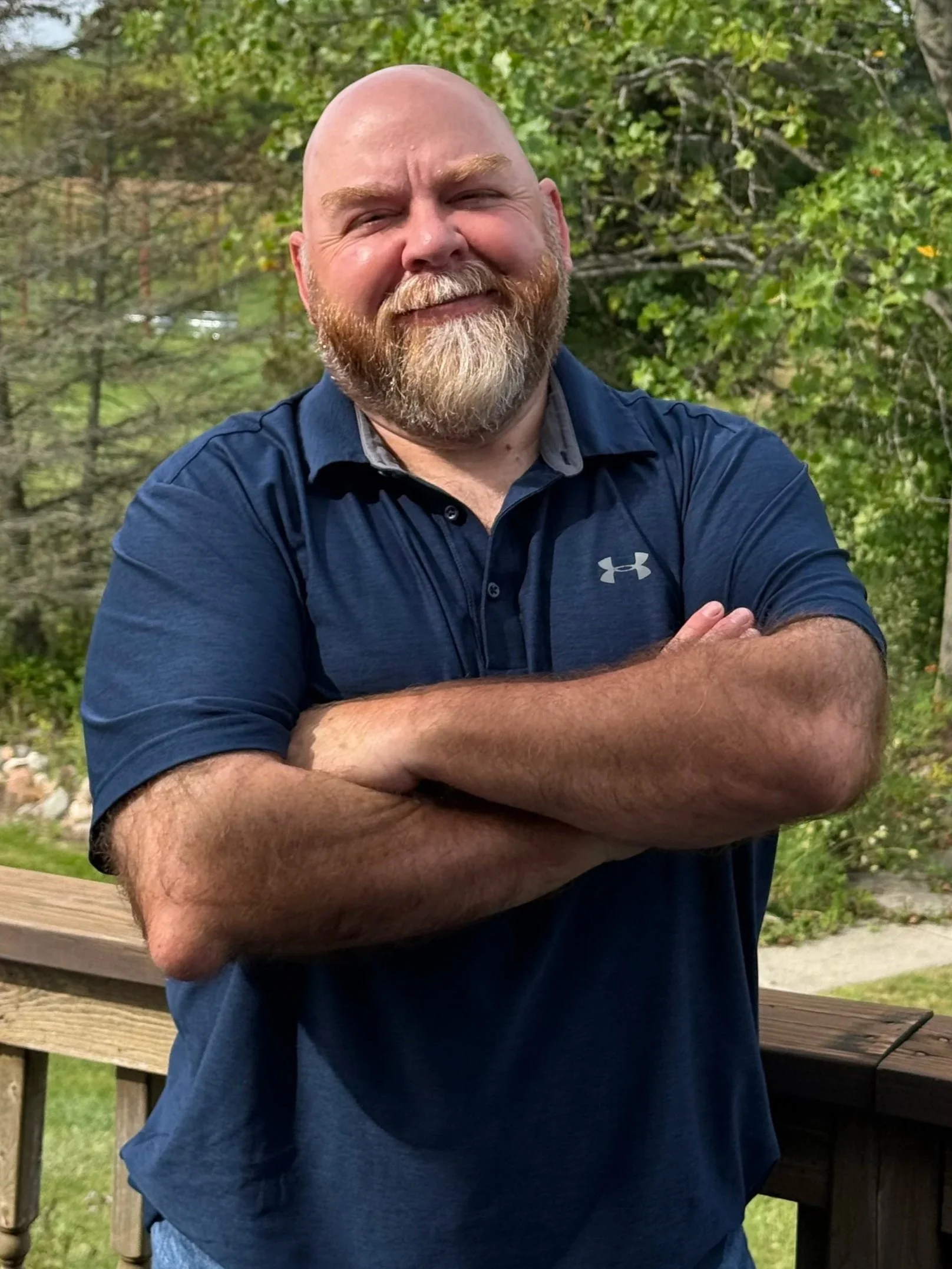 A bald man with a beard standing outdoors in front of trees, crossing his arms and smiling.