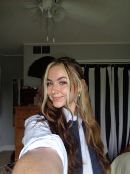 Young woman with wavy, two-toned hair taking a selfie in a room with black and white curtains and a ceiling fan.