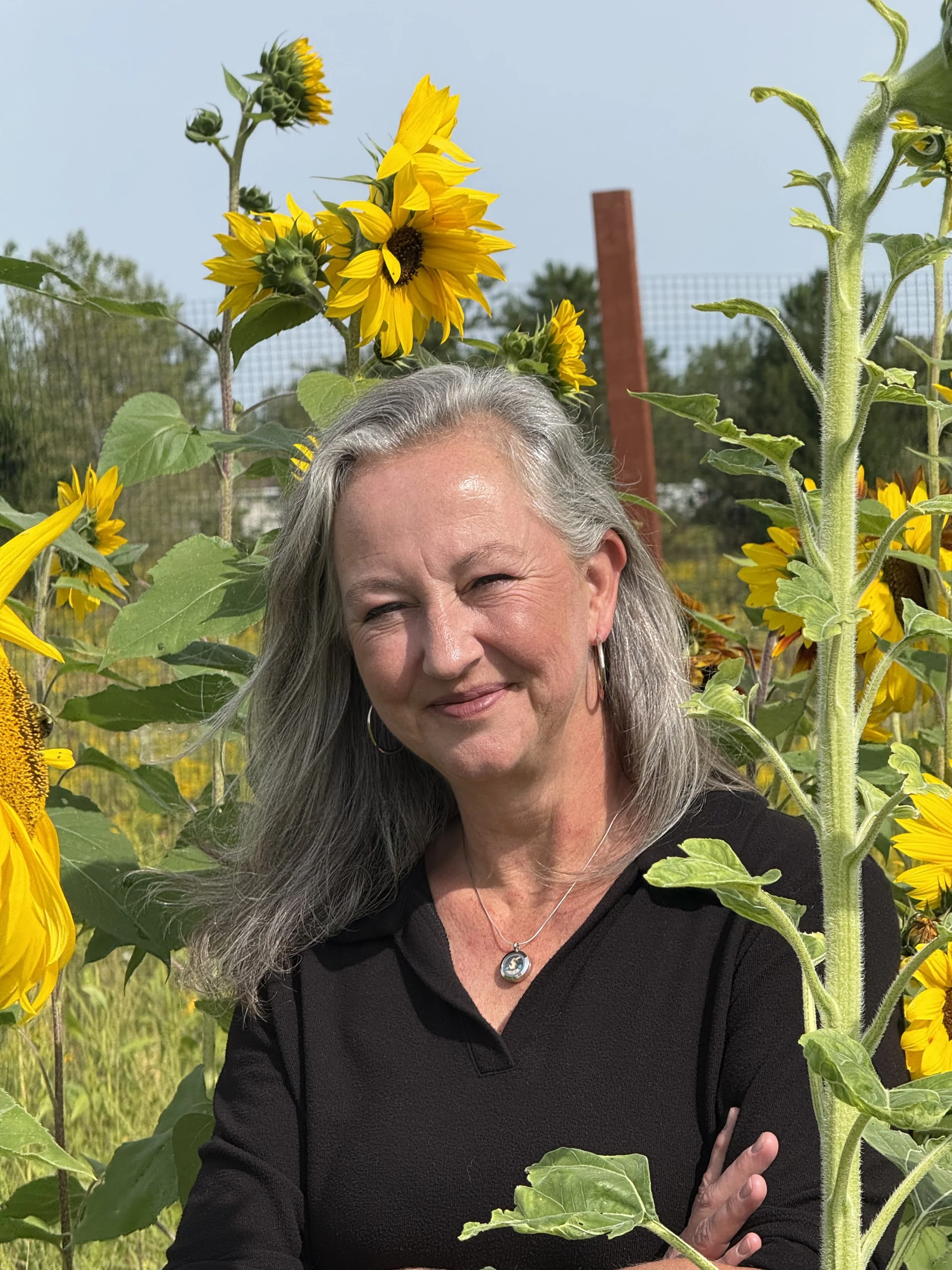A middle-aged woman with gray hair and a necklace featuring a pendant, smiling outdoors among tall sunflowers, with a clear sky and some trees in the background.