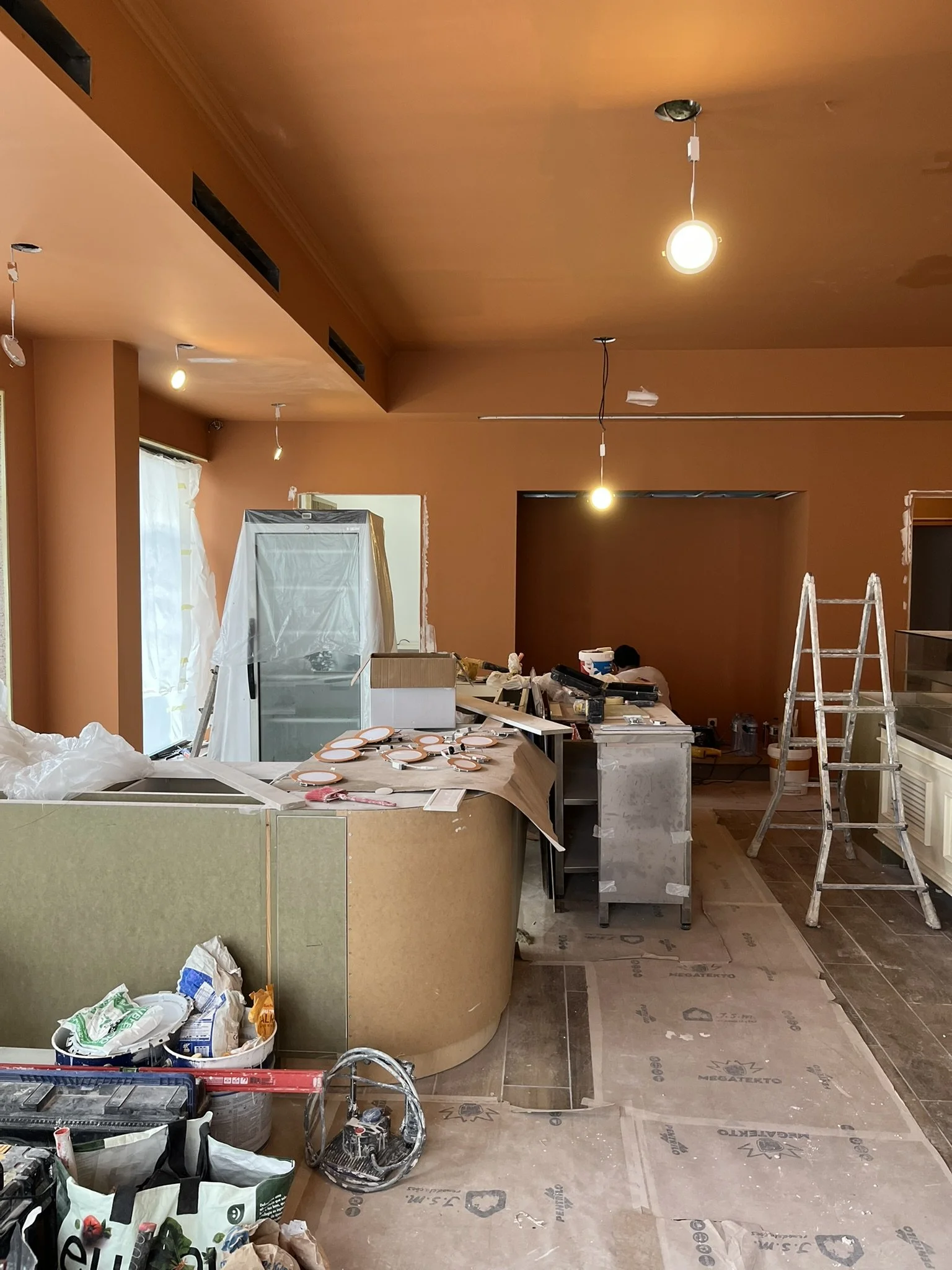Imagem de uma sala em reforma com teto e paredes pintadas de cor laranja, ferramentas, materiais de construção, escadas e objetos dispostos pelo ambiente, indicando trabalho de retrofit ou construção.