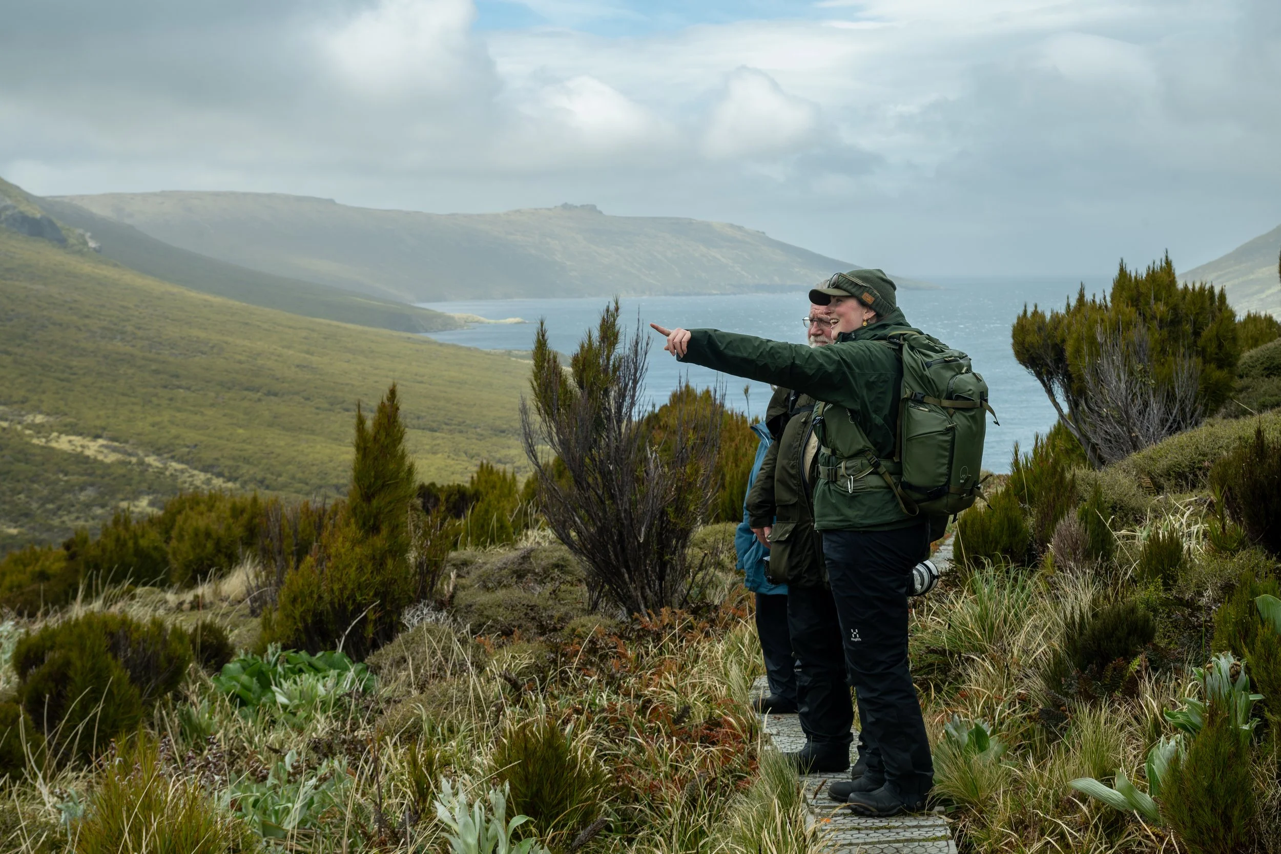 A photograph of Rachel Bigsby pointing toward the distance, beside a workshop guest. They stand on a trail on Campbell Island amidst lush greenery and bushes, with a view of the sea and rolling hills in the background under a partly cloudy sky. 