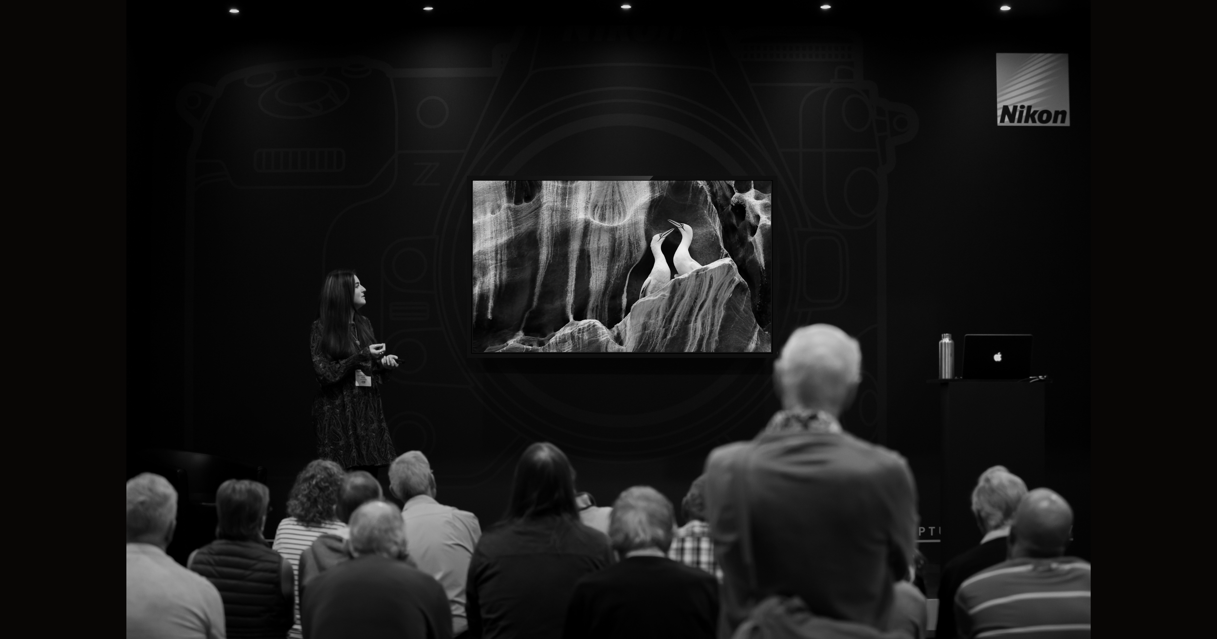 A woman giving a presentation on photography at a Nikon event. She holds a remote control and stands in front of a screen showing a black and white photo of two birds standing on rocks in a canyon. Audience members are seated and watching her.