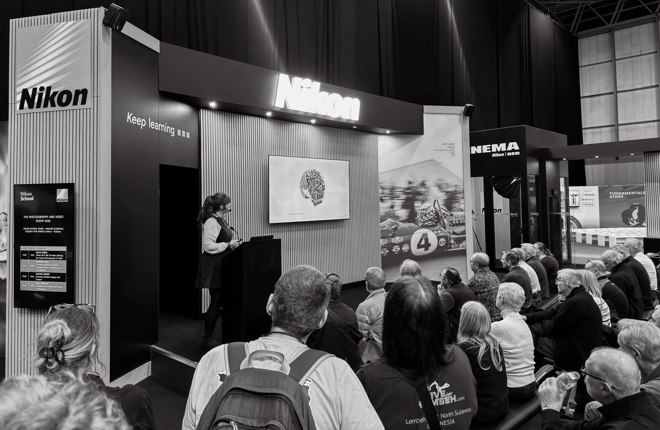 A black and white photo of a Nikon booth at a photography event, with a speaker on stage and an audience of seated and standing people listening.