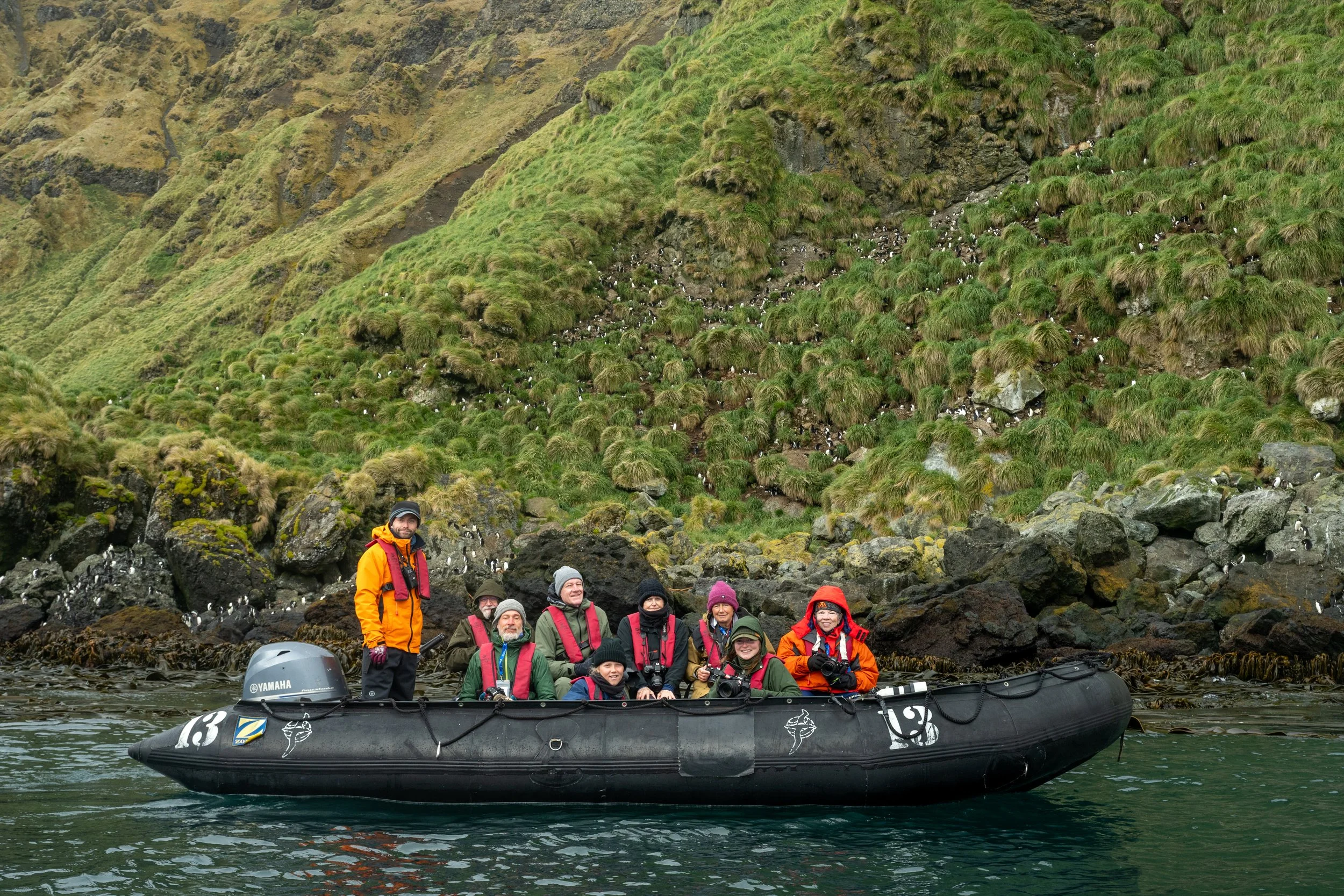 A photograph of workshop guests in a black inflatable boat on the Southern Ocean waters surrounding Macquarie Island, with green, rocky cliffs and many seabirds in the background.