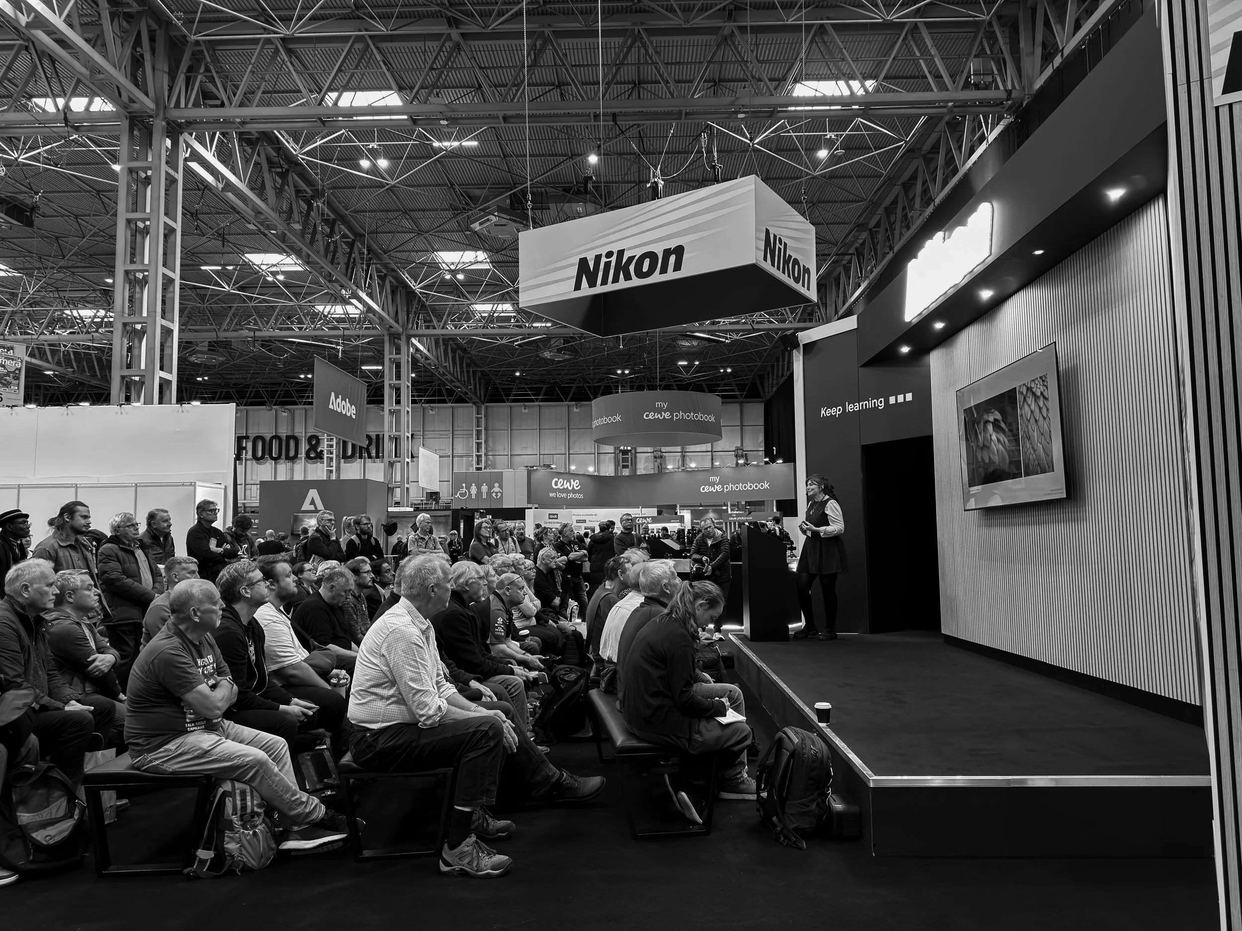 A large crowd of people seated in front of a stage at a convention or conference, with Nikon and Adobe banners hanging overhead in a spacious exhibition hall.