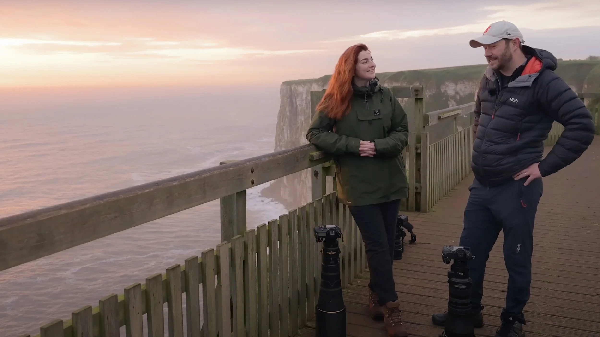 A photograph of Rachel Bigsby stood beside a workshop guest on a wooden viewing platform near at RSPB Bempton Cliffs, smiling and chatting during sunrise, with cameras on tripods nearby.