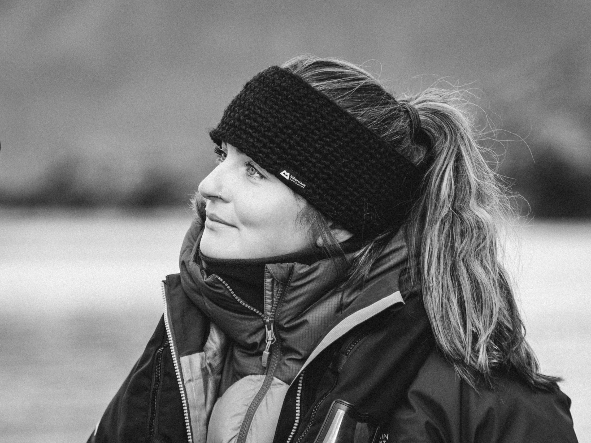 A black and white photograph of Rachel Bigsby wearing a knitted headband and a winter jacket, looking to her right with snowy landscape of Arctic Svalbard in the background.