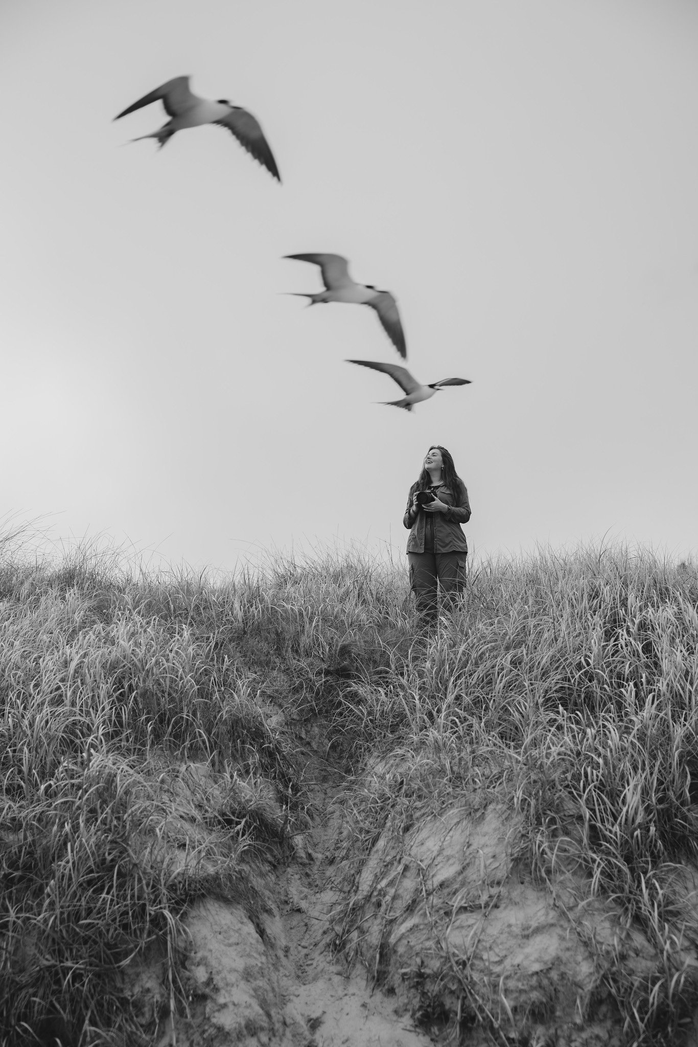 A black and white portrait of Rachel Bigsby, standing on a grassy sand dune on Lord Howe Island, Australia, holding a Nikon camera, looking up at Sooty Terns flying in the muted sky over her.