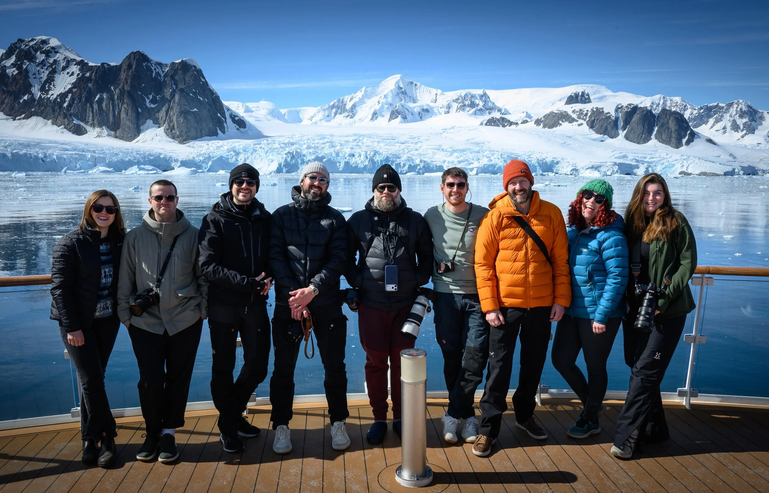 A photograph of a group of nine people (including Rachel Bigsby on the farthest right) standing on the deck of a boat in Antarctica with snowy mountains and ice in the background, wearing winter clothing and sunglasses.