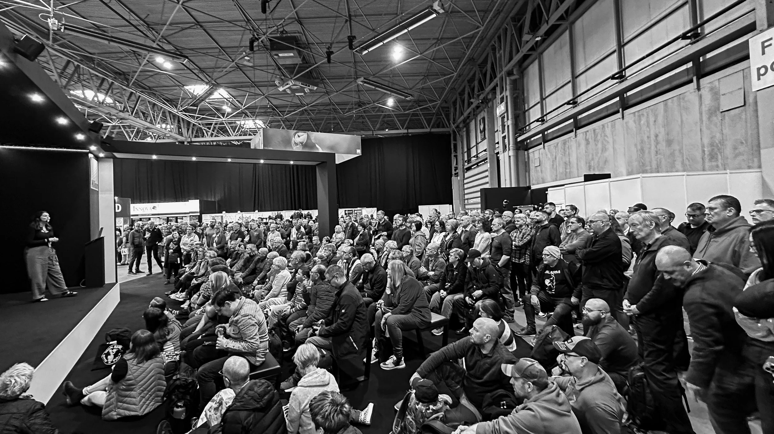 A black and white photograph of Rachel Bigsby standing onstage, mid-speech, presenting a wildlife photography lecture as a large crowd watch.