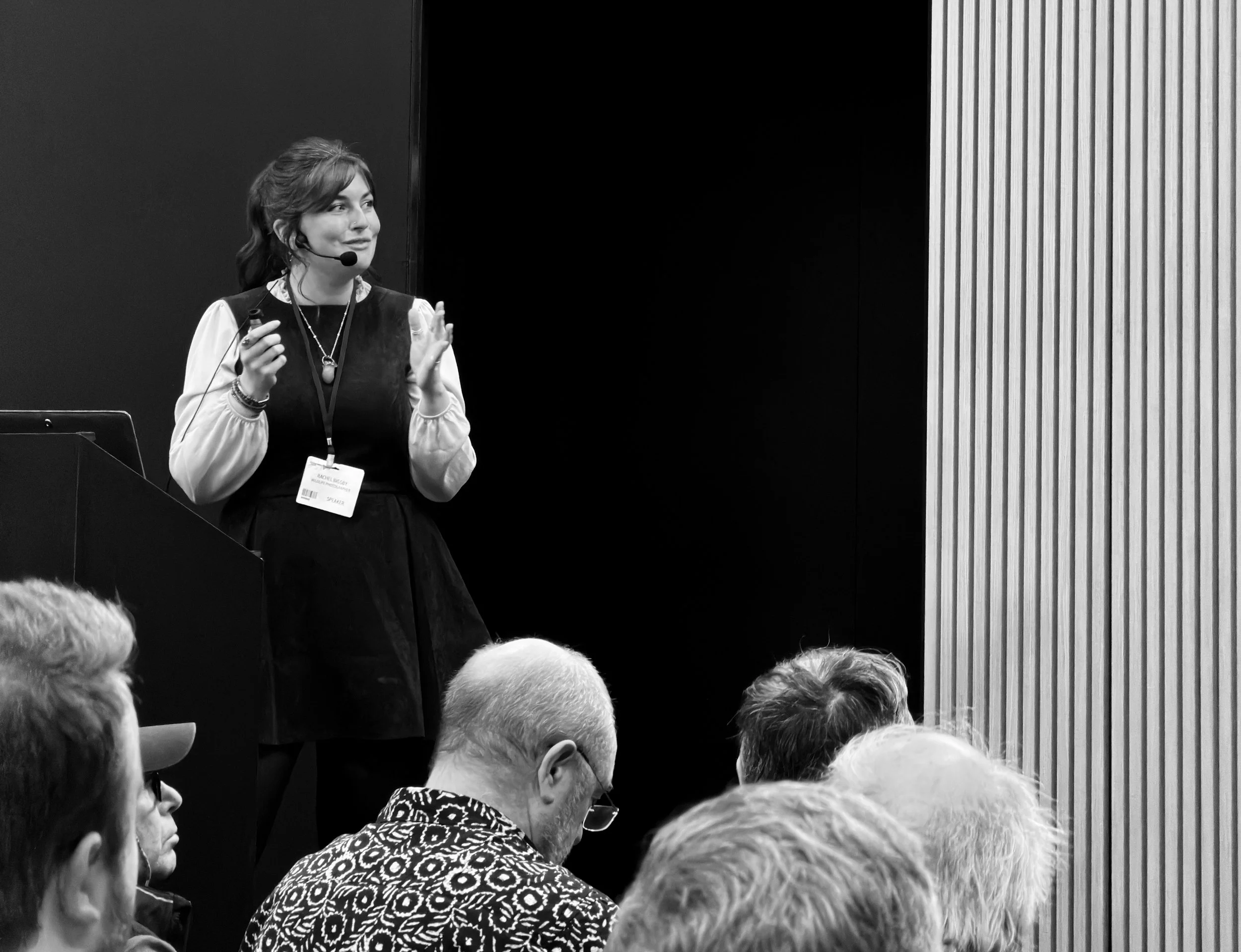 A black and white photograph of Rachel Bigsby standing onstage, mid-speech, presenting a wildlife photography lecture.