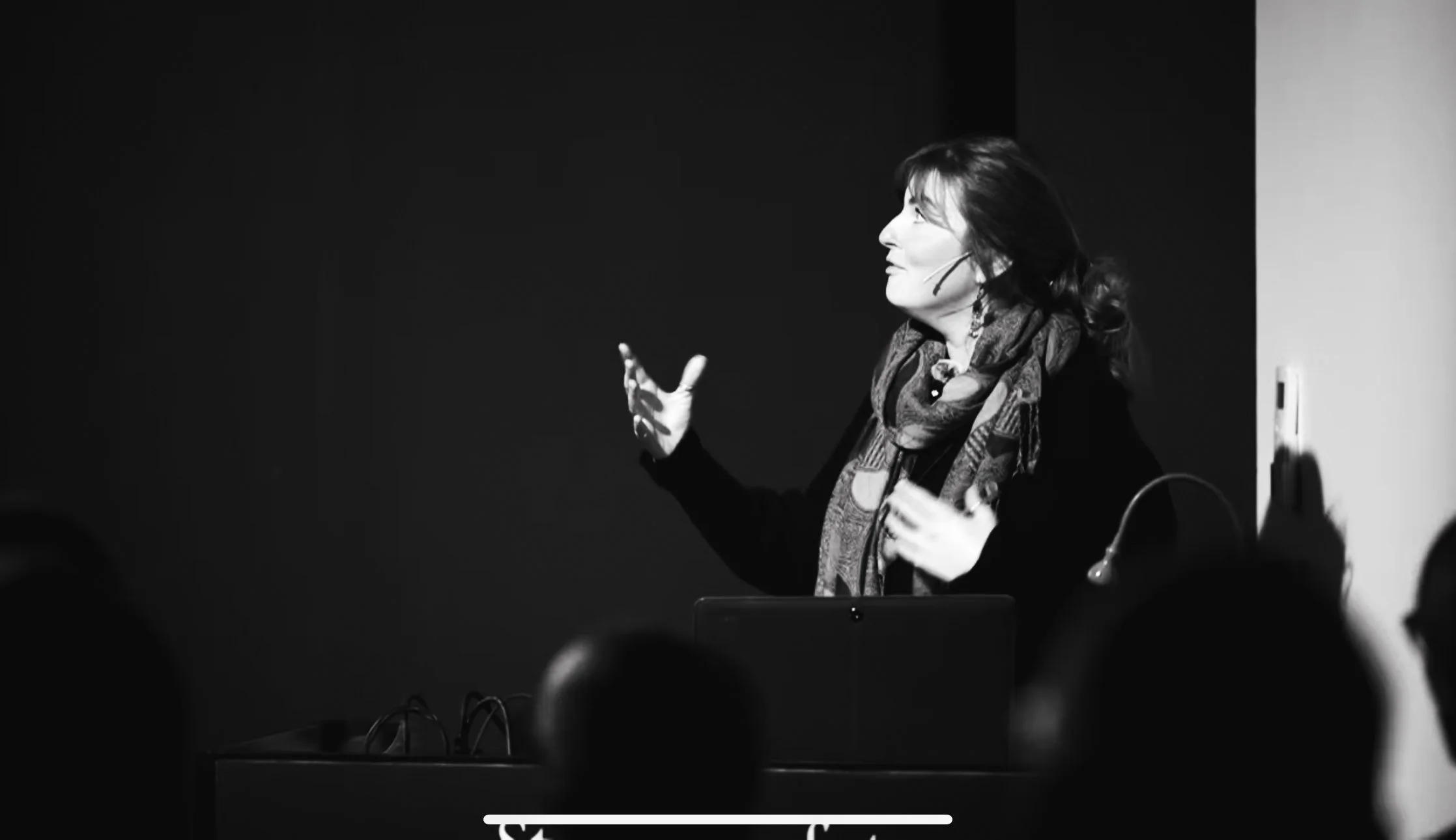 A woman speaking or presenting in front of a dark background with a black and white photo, wearing a patterned scarf and jacket, using a headset microphone.