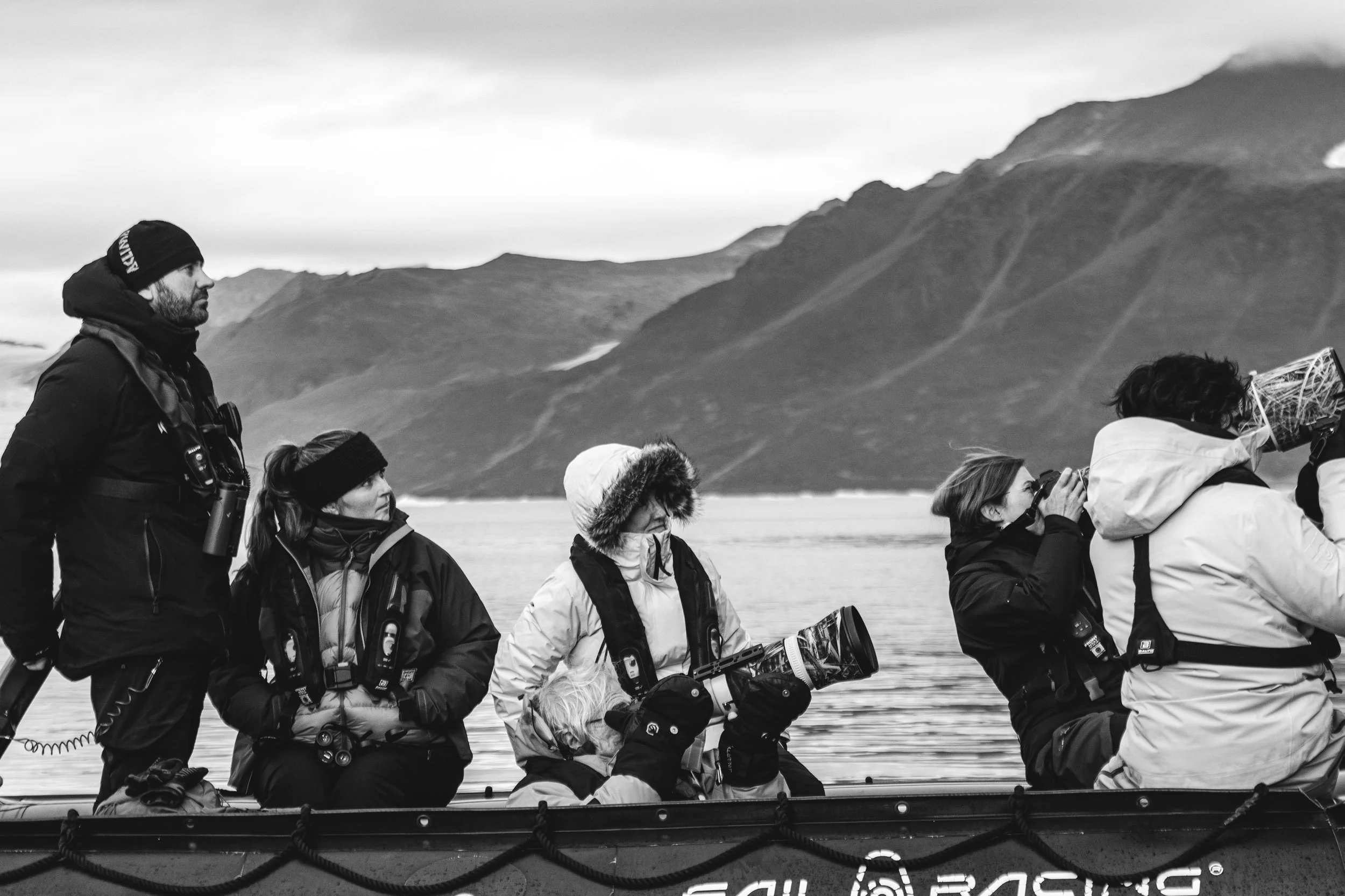 A black and white photograph of Rachel Bigsby's workshop guests in outdoor winter clothing on a boat in Arctic Svalbard, with mountains in the background.