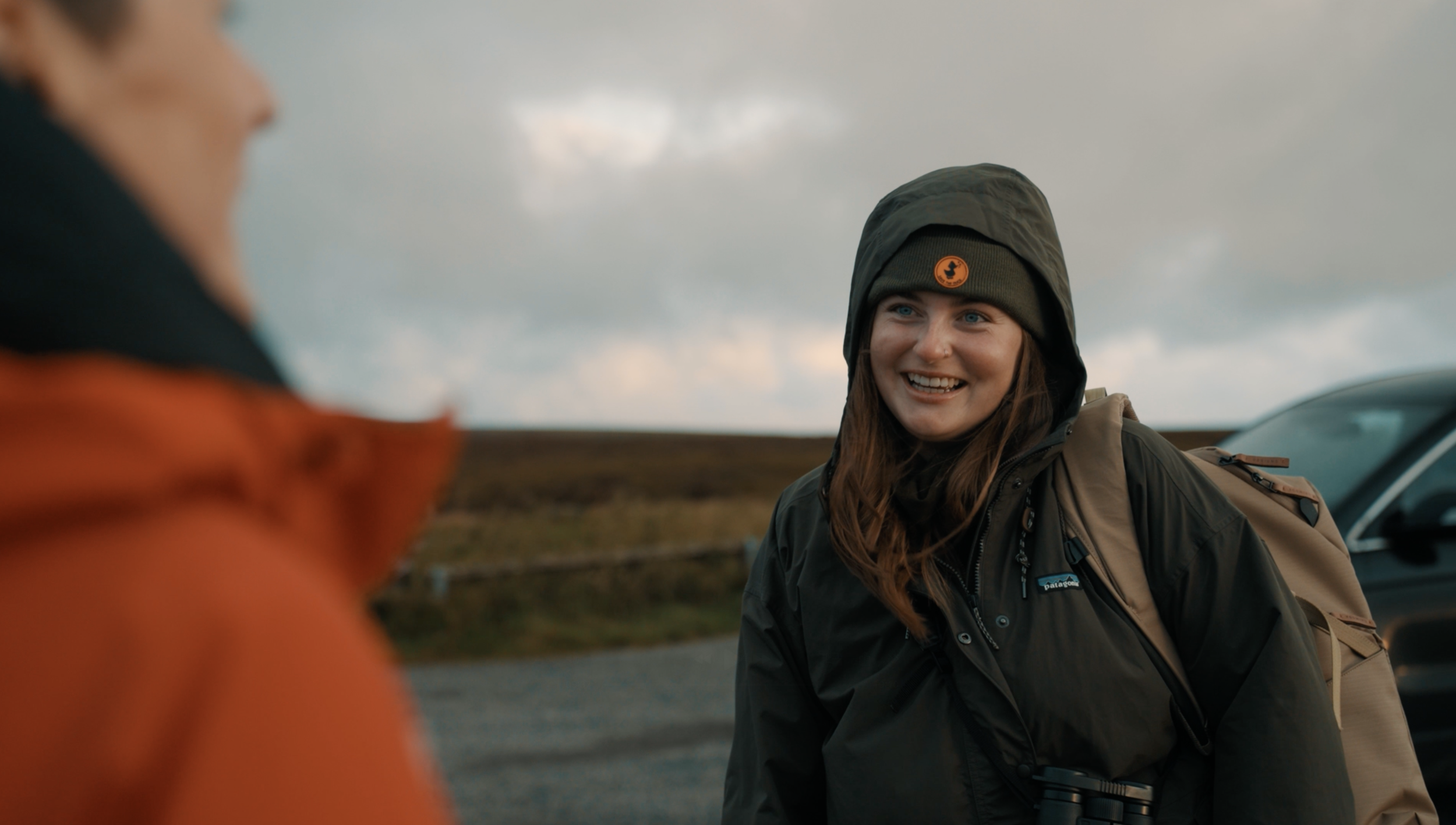A photograph of Rachel Bigsby smiling at a workshop guest, wearing a green beanie and outdoor jacket, outdoors with cloudy sky in the background.