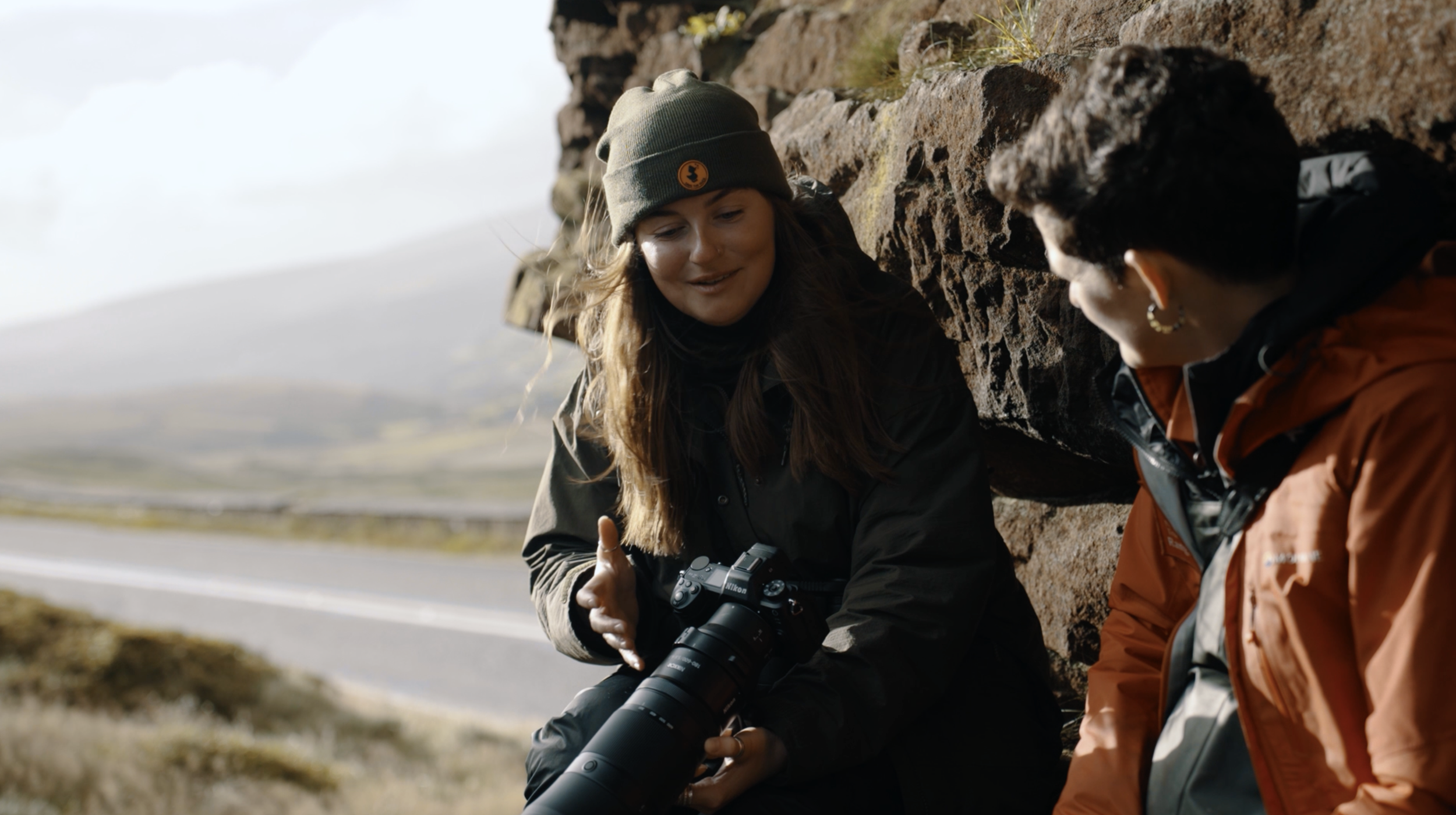A photograph of Rachel Bigsby sitting beside a workshop guest outdoors near a rocky area, showing the guest a Nikon camera, in soft Autumnal light.