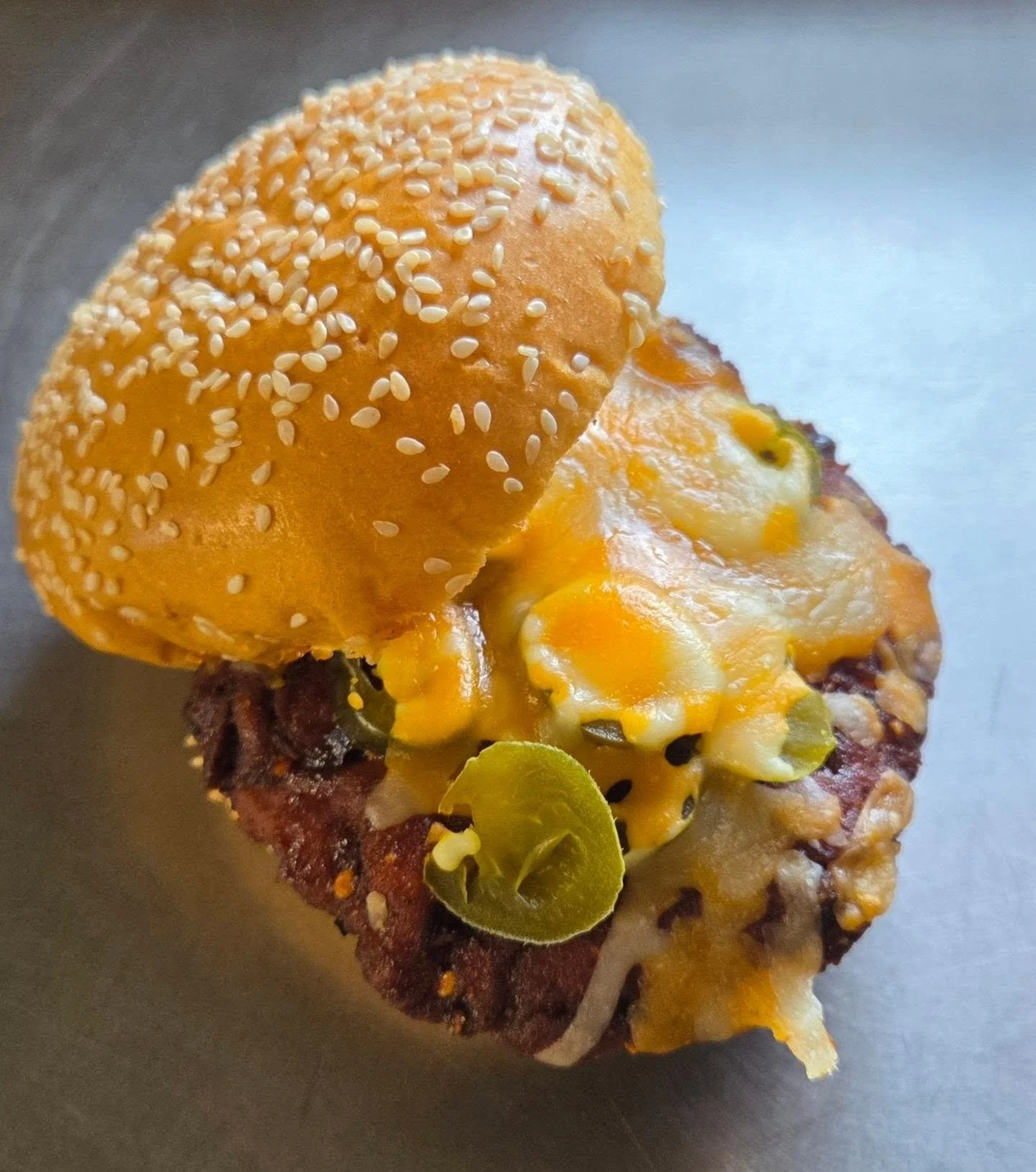 Close-up of a breakfast dish featuring a biscuit topped with melted cheese, a slice of jalapeño, and crispy bacon on a dark surface.