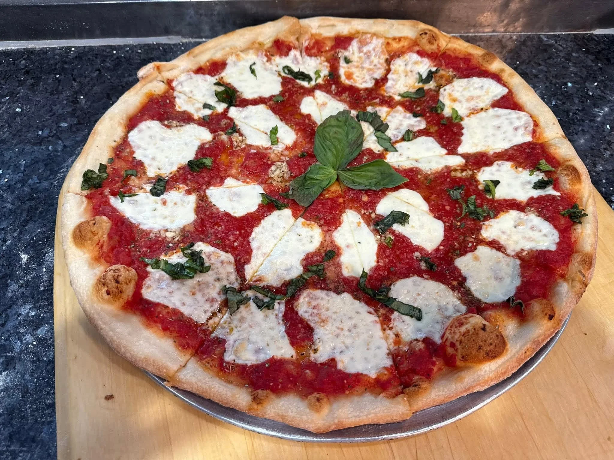 A pizza with a golden crust topped with tomato sauce, slices of mozzarella cheese, chopped basil, and fresh basil leaves in the center. The pizza is on a round metal tray placed on a black speckled countertop.