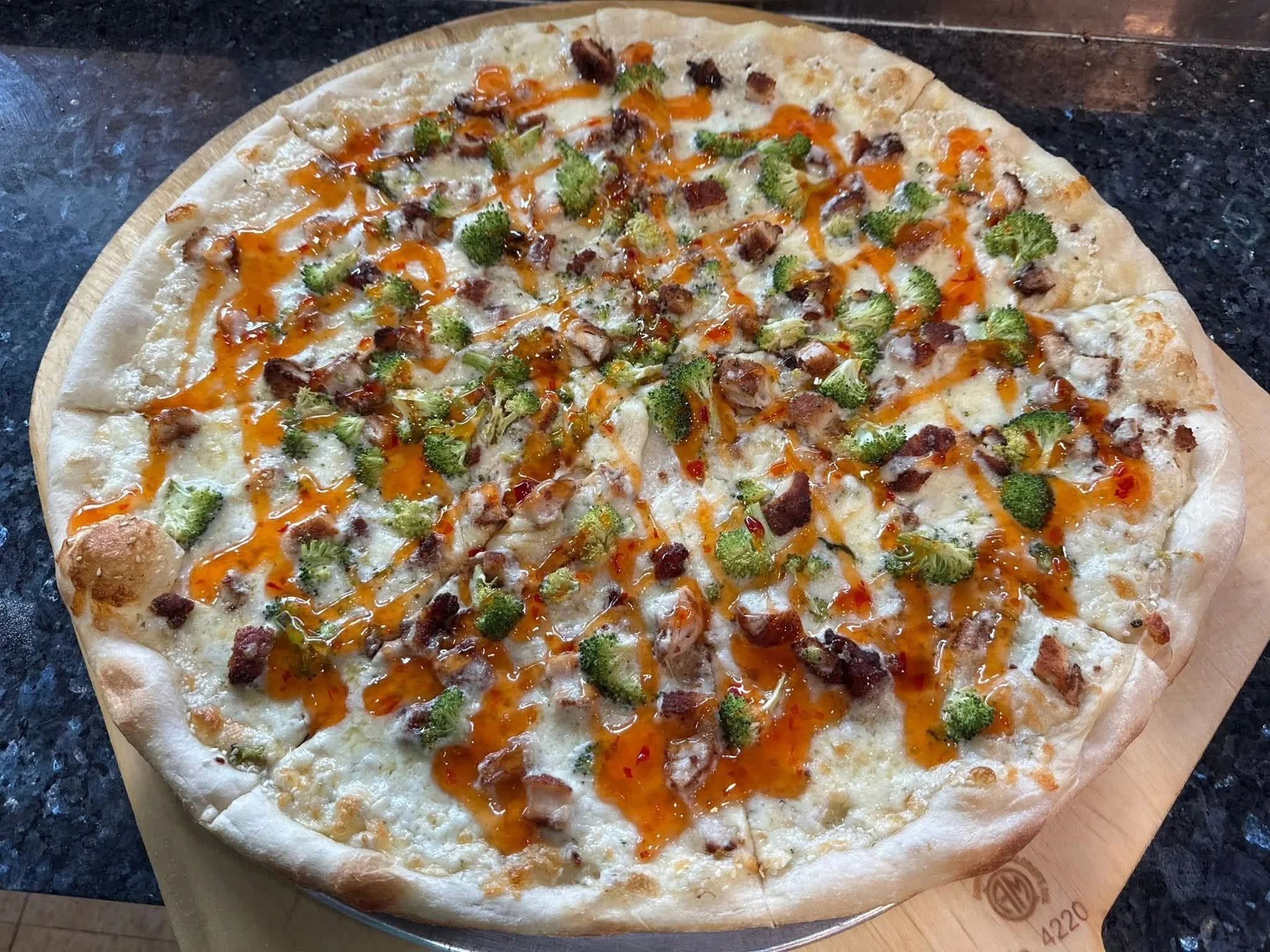 A pizza with a thick crust topped with cheese, broccoli, and pieces of meat or bacon, drizzled with orange-colored sauce, on a wooden pizza tray.