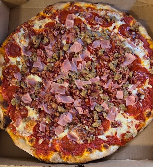 Pizza topped with pepperoni, sausage, bacon bits, and pink ham slices inside a cardboard box.