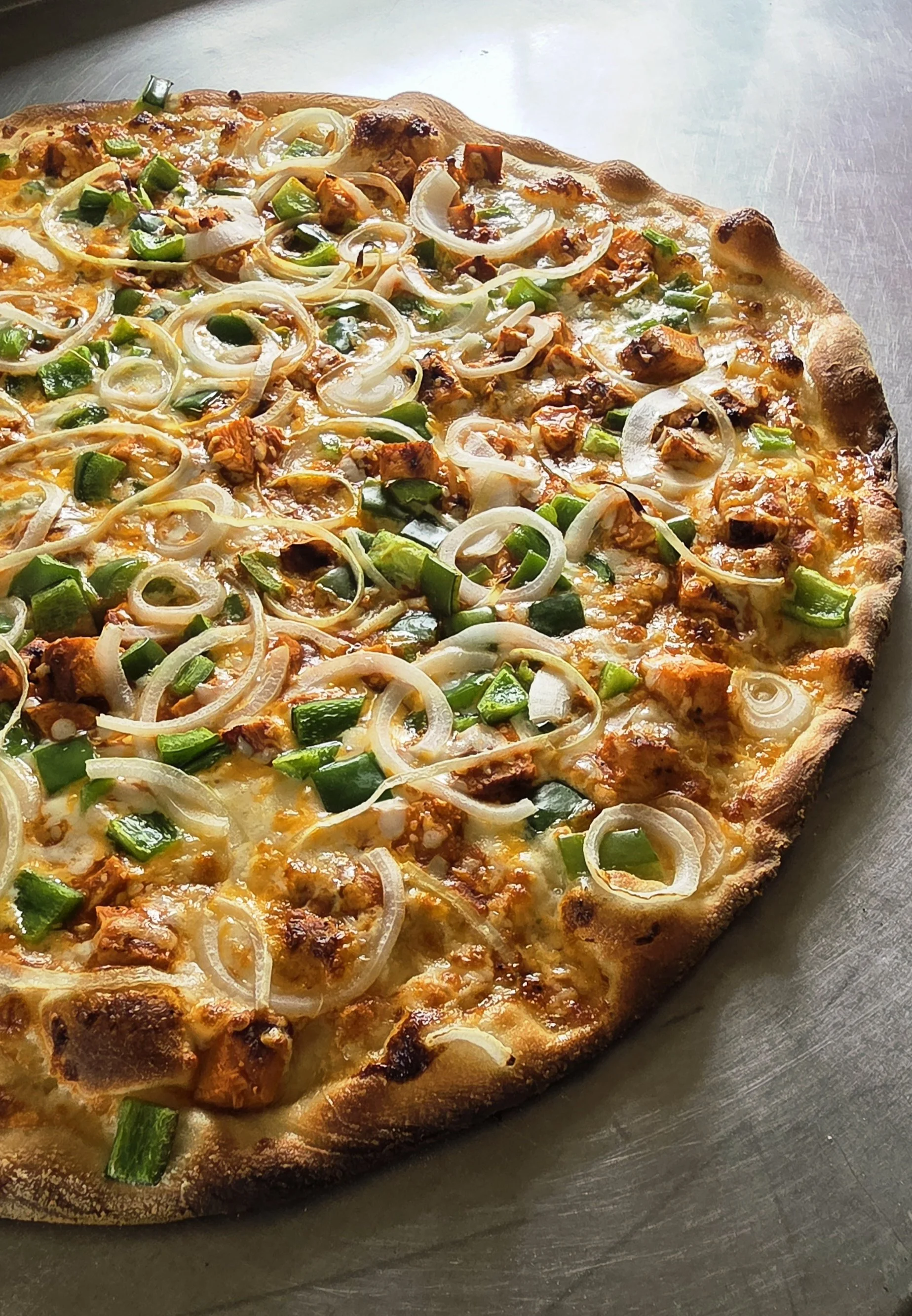 Close-up of a cooked pizza with onion, green pepper, and cubed chicken toppings on a metal surface.