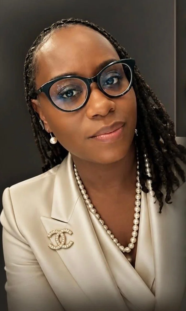 A woman with braids wearing black glasses, pearl jewelry, a beige blazer with a Chanel brooch, and creative eye makeup.