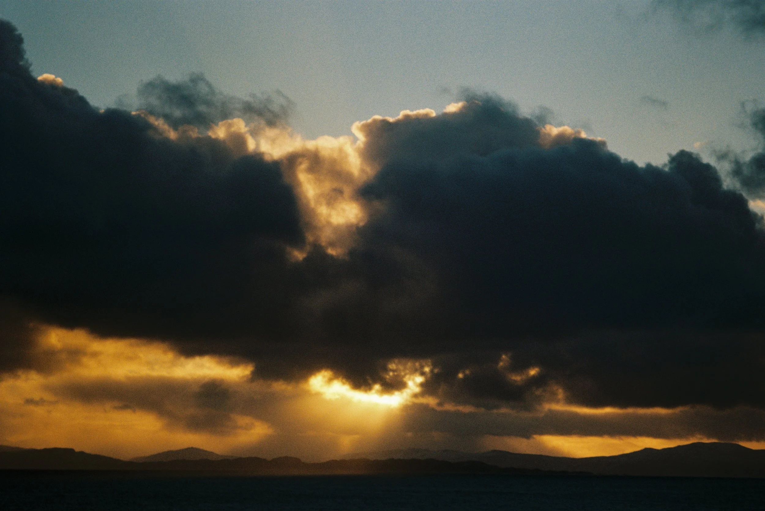 Sunset over the ocean with dark clouds and distant mountains.
