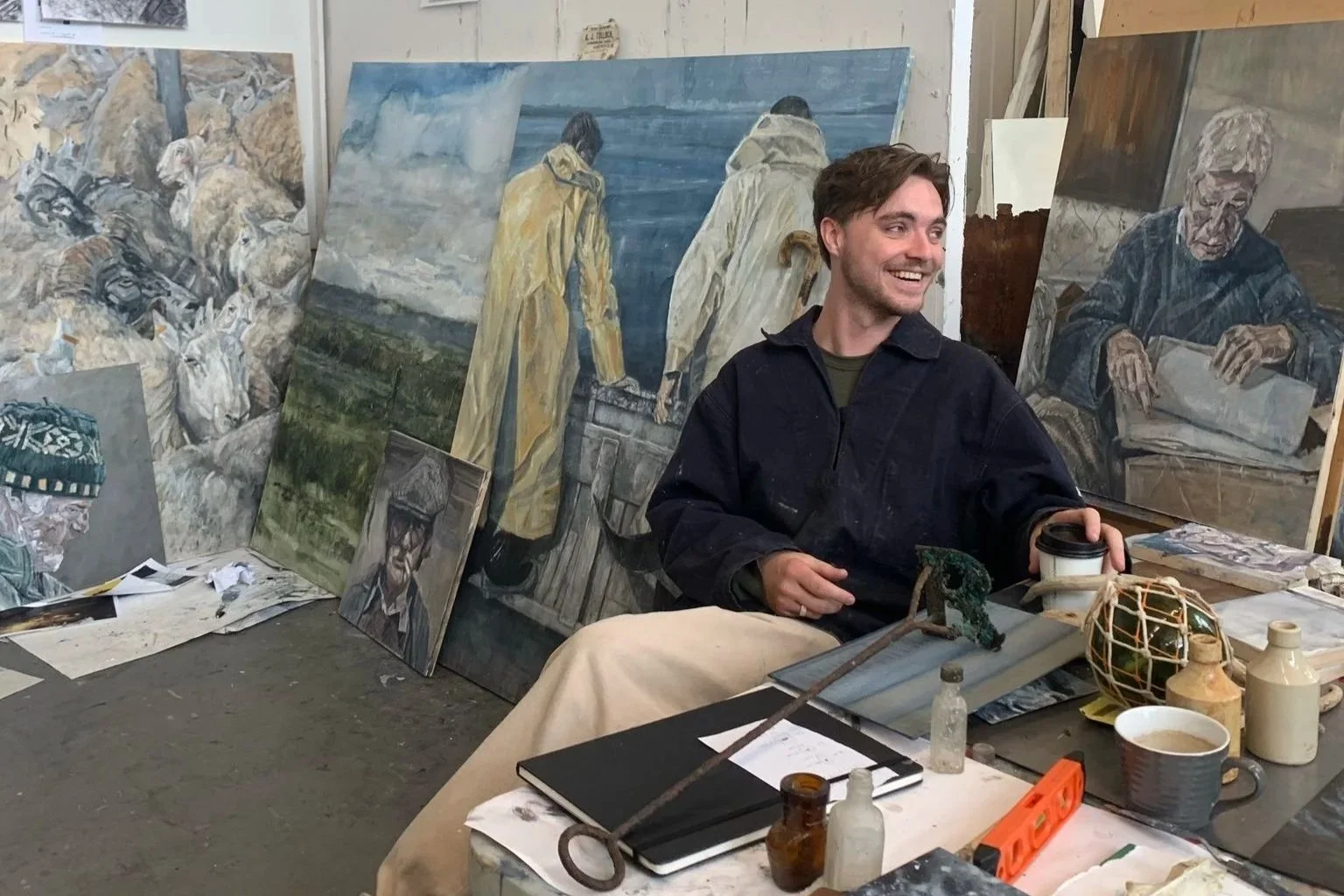 A man sitting at a cluttered table surrounded by several landscape and portrait paintings in an art studio. He is smiling and holding a disposable coffee cup.