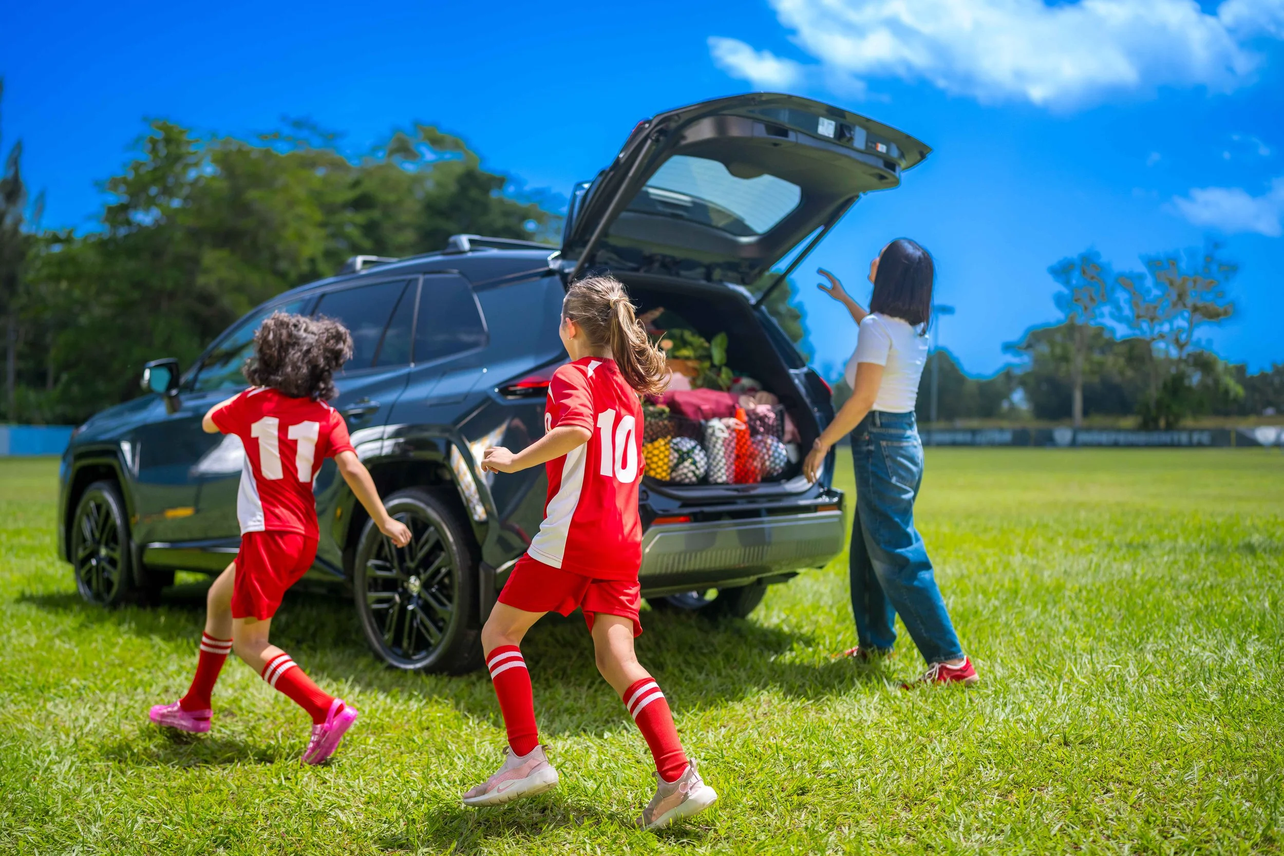 Two children in red soccer uniforms running toward a Toyota RAV4 with its cargo area open beside an adult.