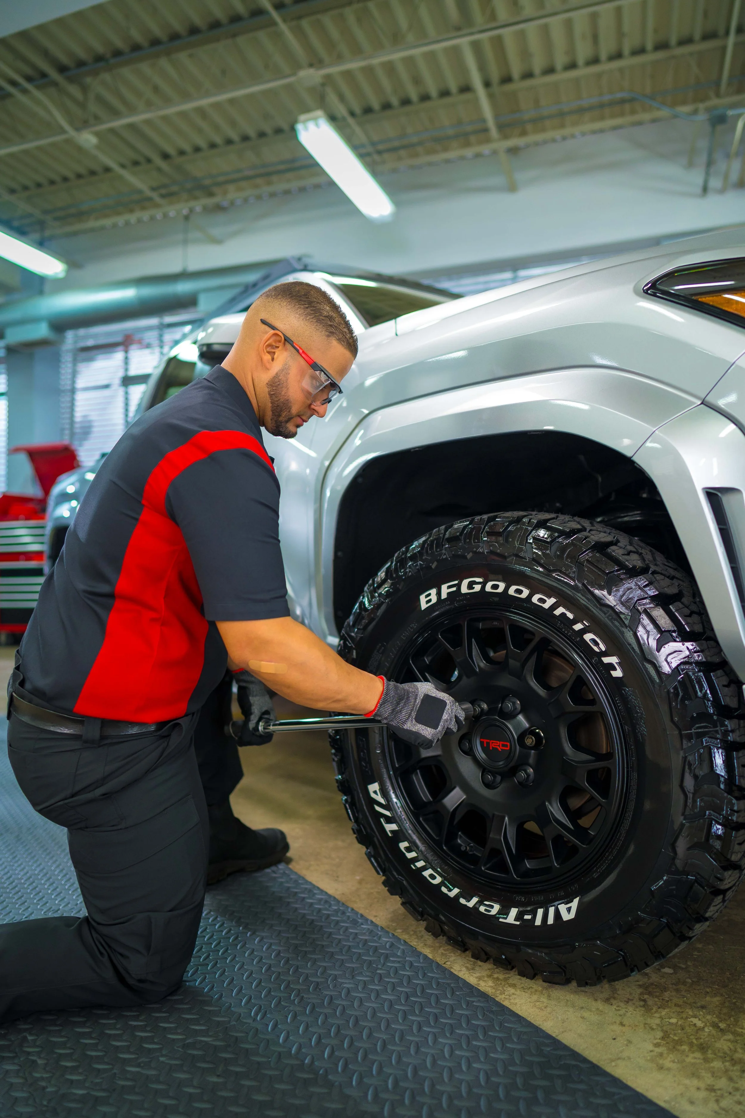 Toyota technician installing a black TRD wheel on a silver Tacoma inside a service shop.