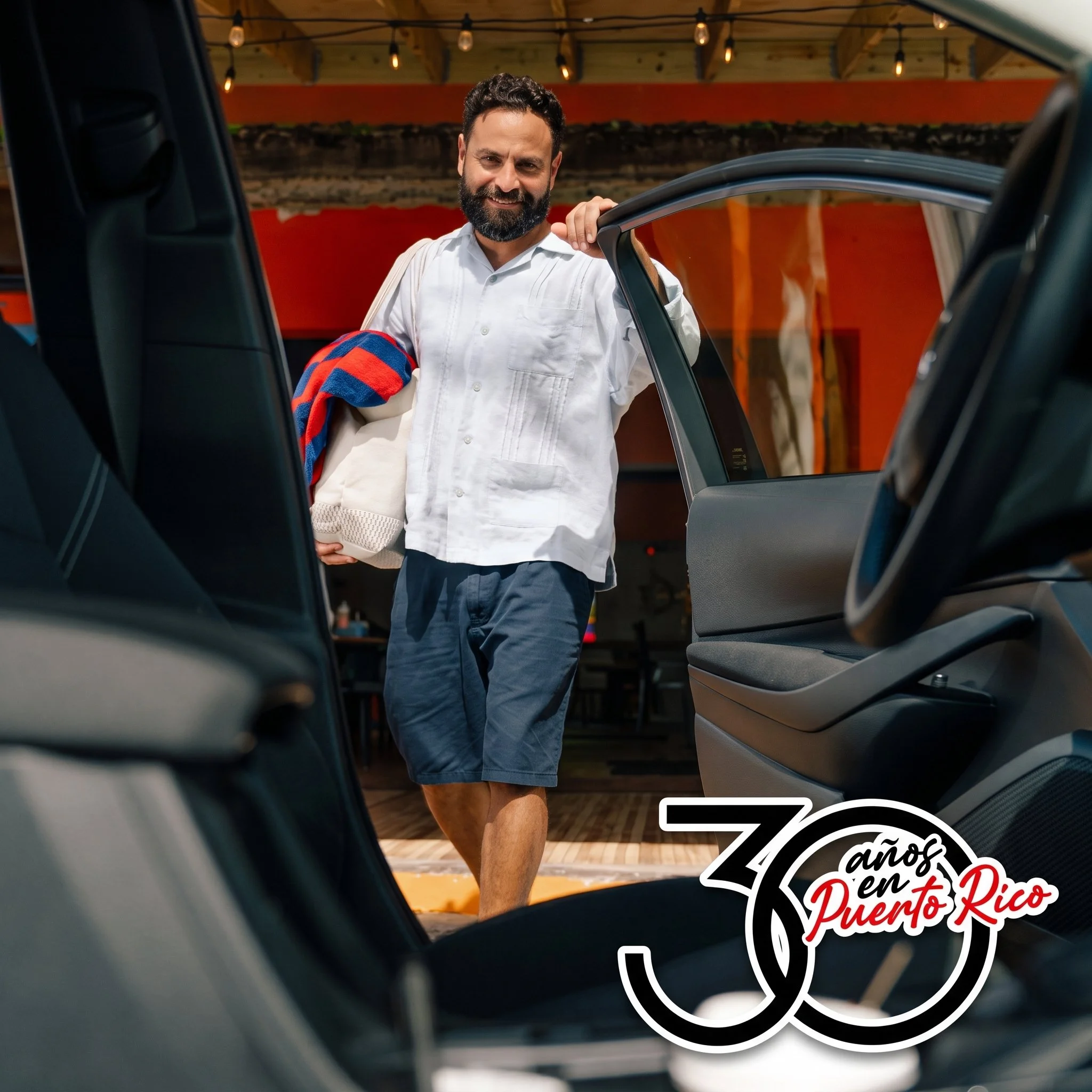 Person standing beside an open vehicle door holding beach gear, with the “30 años en Puerto Rico” anniversary graphic.