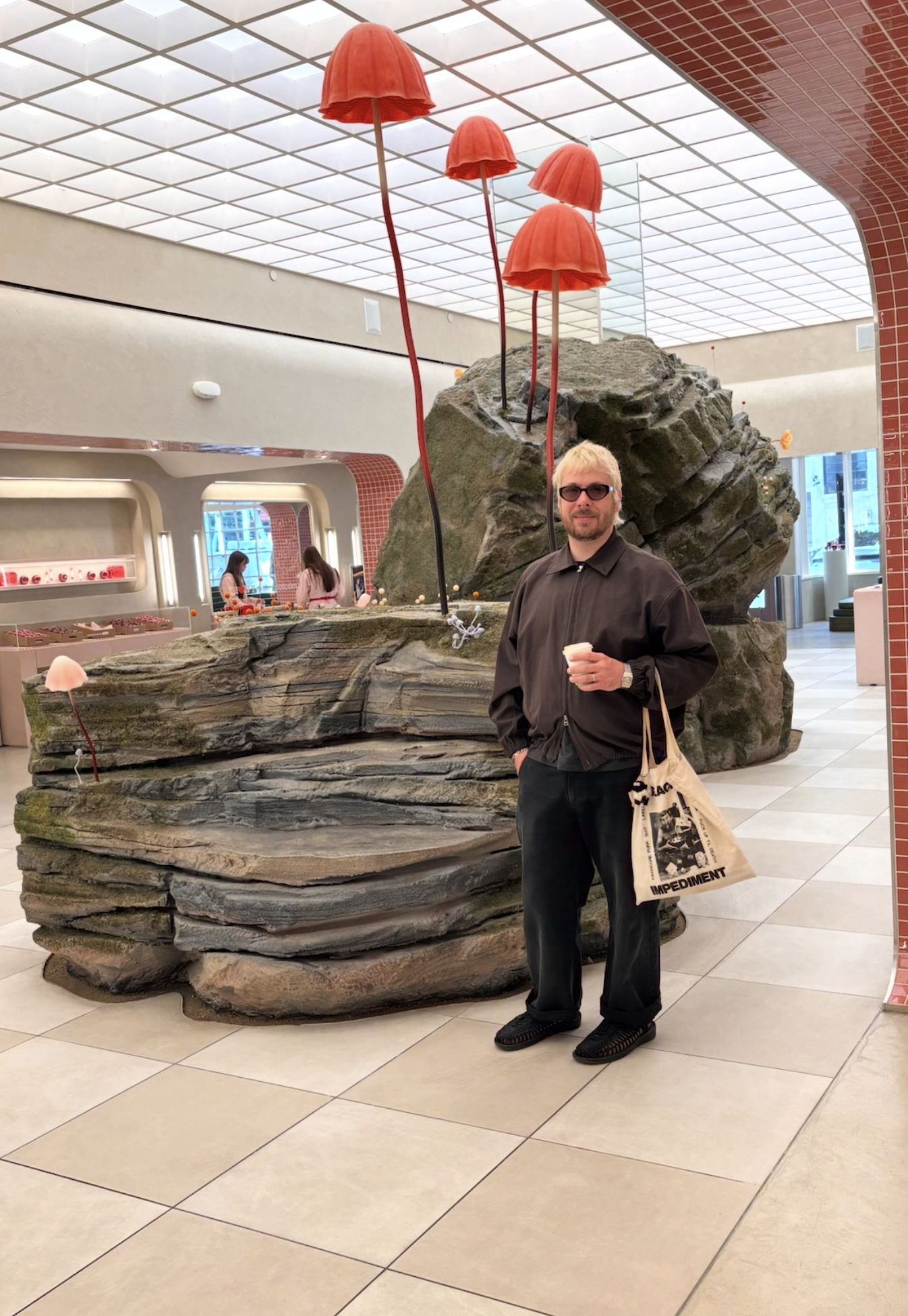 A man wearing sunglasses and black clothing standing in front of a large, rock-like sculpture with red umbrella-like structures on metal poles. The man is holding a cup and a beige tote bag with black text and images.