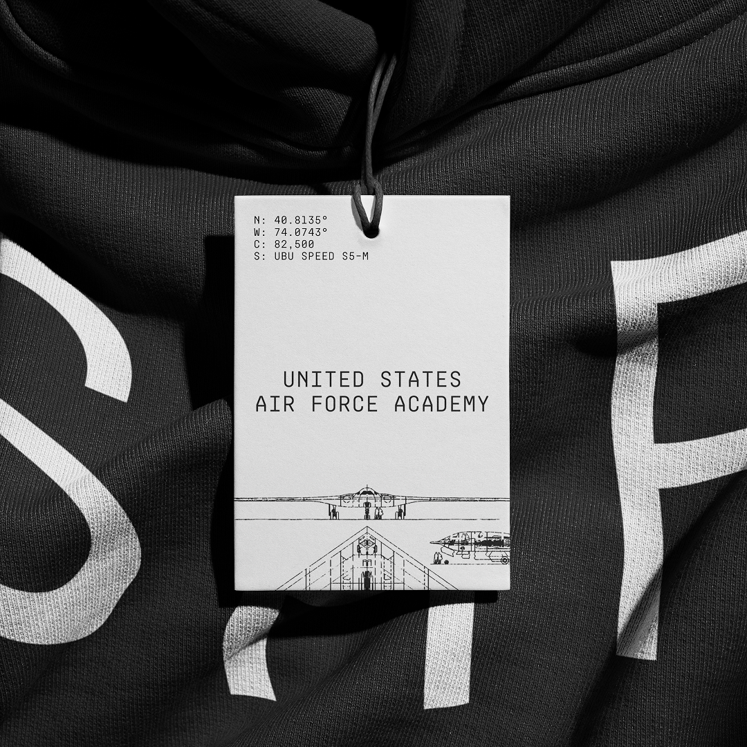 Binocular flight illustration on a tag reads 'United States Air Force Academy' and includes geographical coordinates and aircraft specifications, attached to black fabric with white lettering.