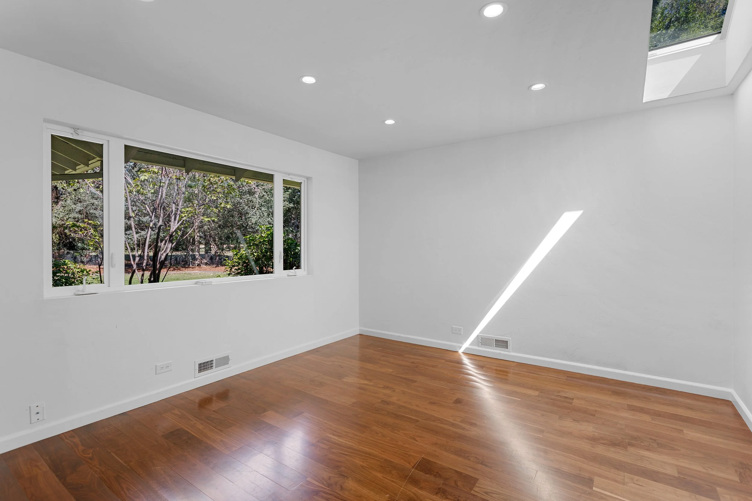 Empty room with white walls, hardwood flooring, a large window showing trees outside, ceiling lights, and a skylight.