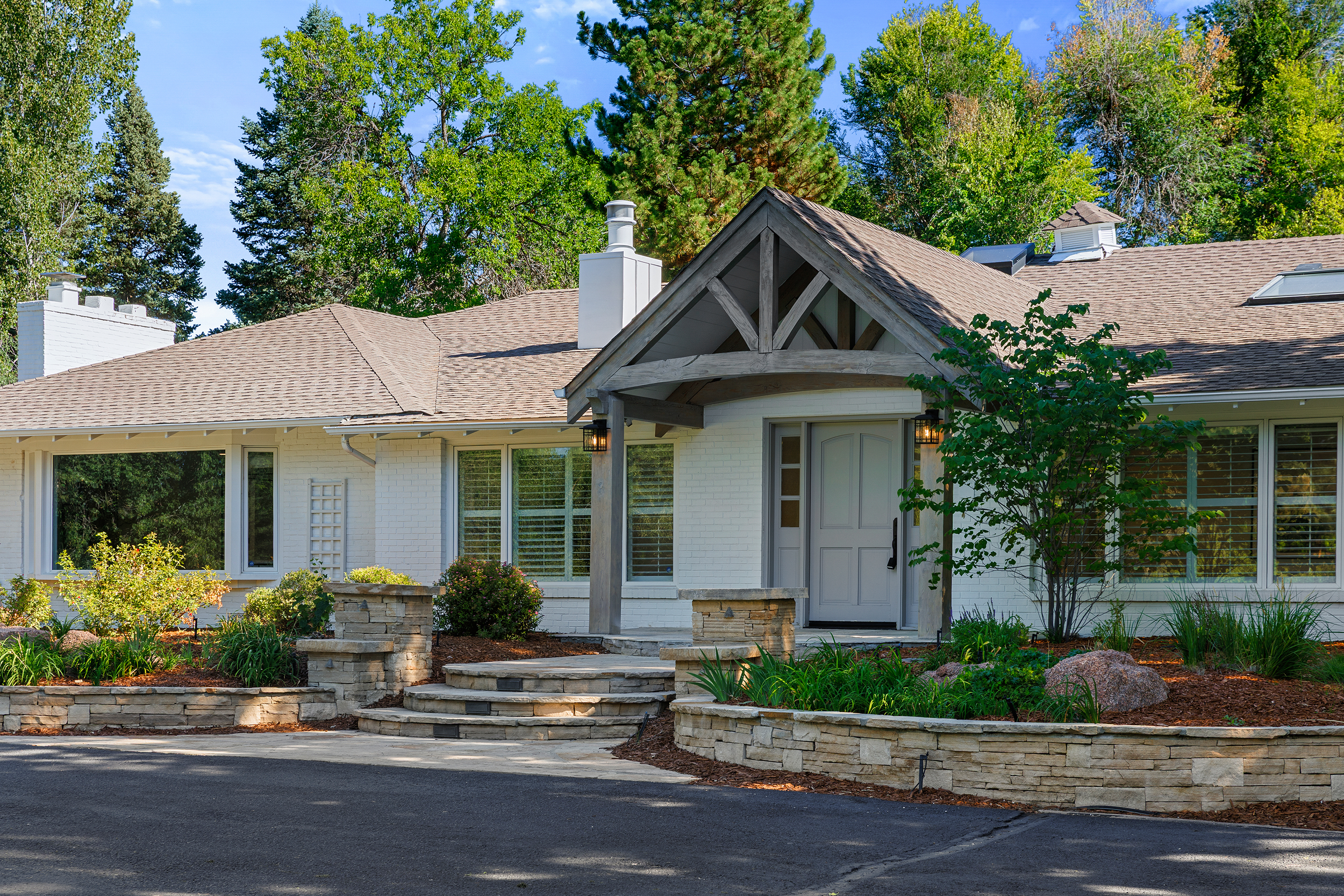 Front view of a white brick house with a gabled roof, surrounded by lush green trees and a landscaped garden with stone steps and a curved stone wall.