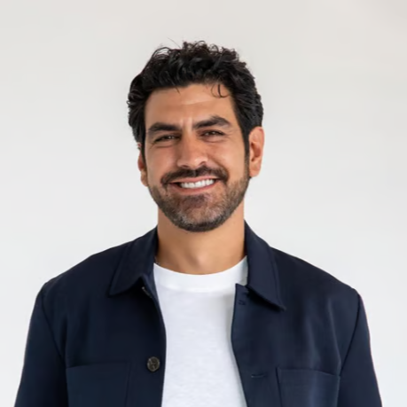 Smiling man with dark curly hair and beard wearing a white shirt and navy jacket against a plain light gray background.