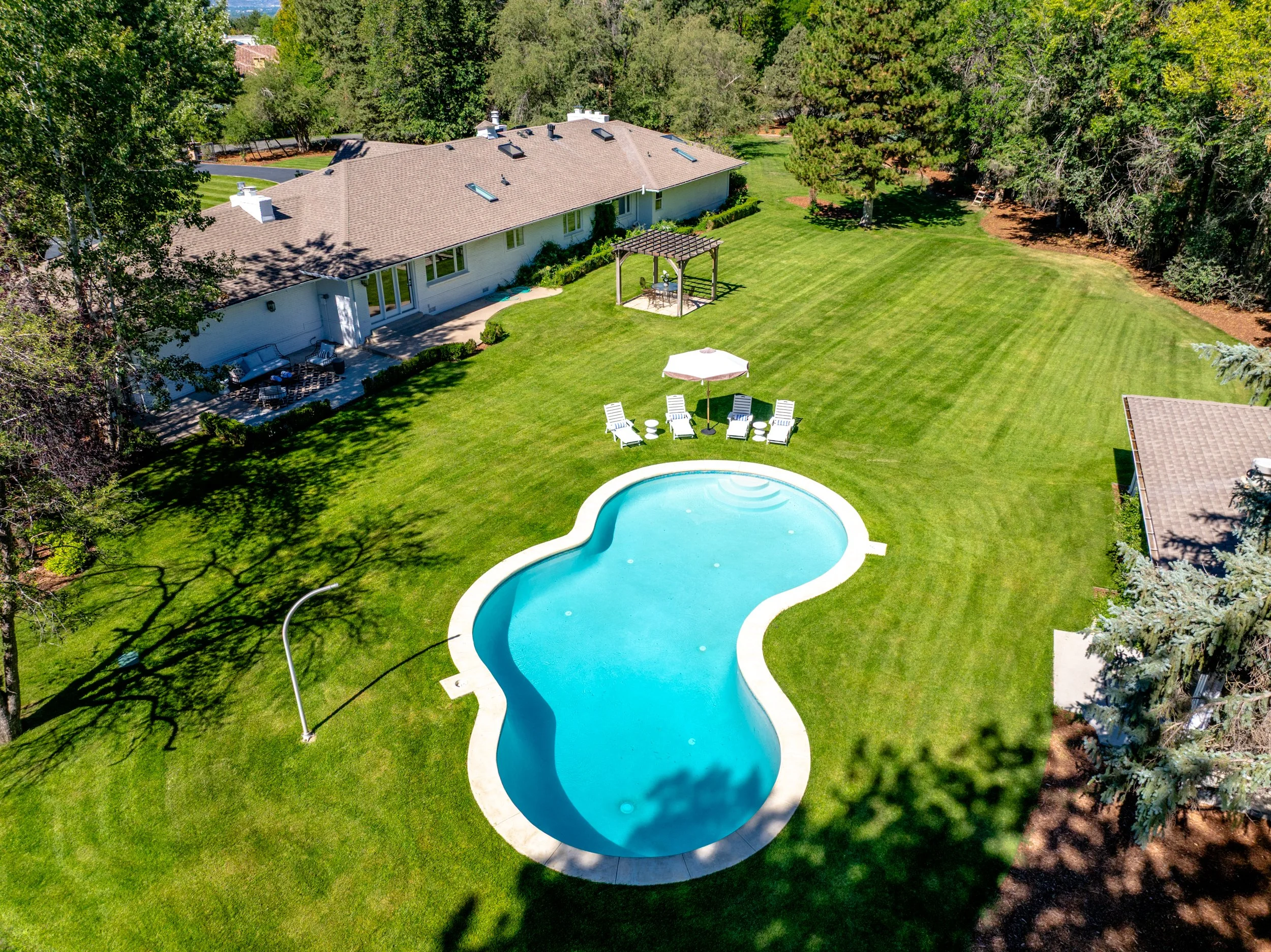 Aerial view of a backyard with a swimming pool, green lawn, lounge chairs with an umbrella, a patio area with outdoor furniture, and a covered seating area with a pergola, surrounded by trees.