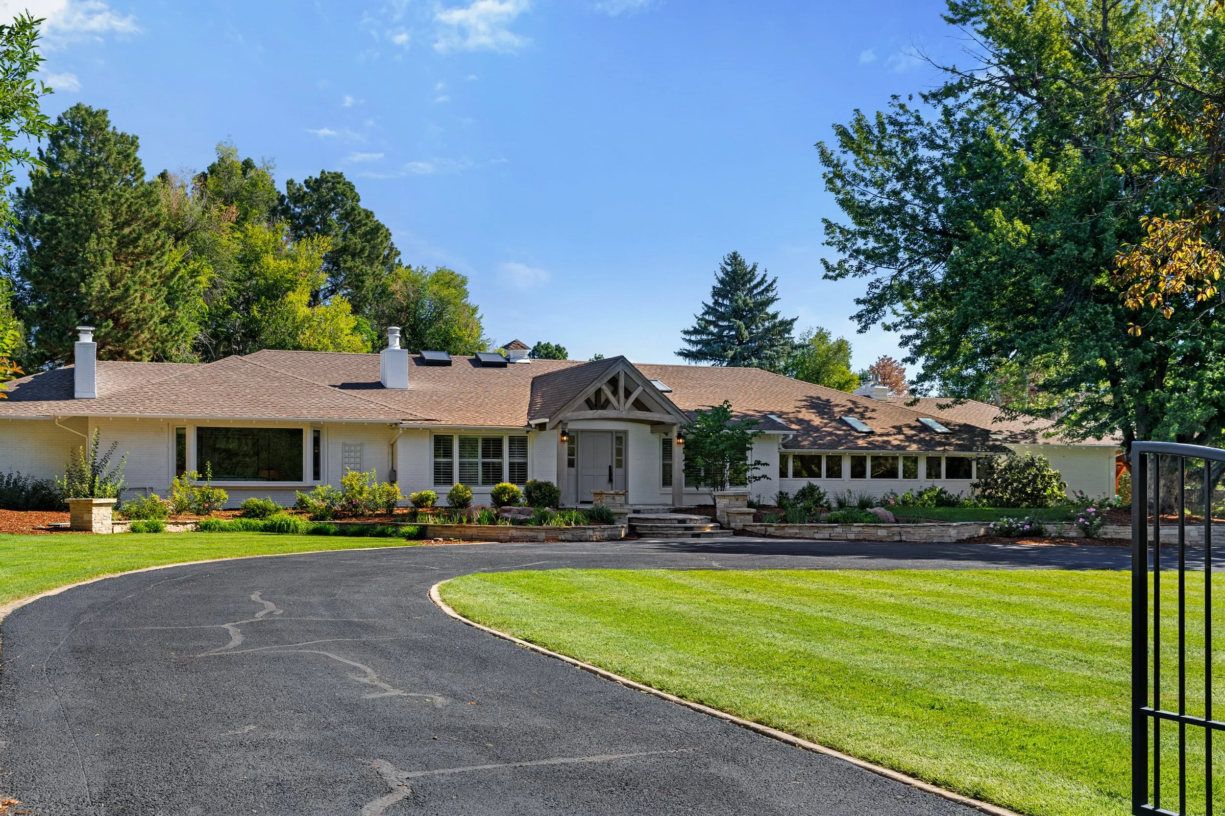 A house with a brown roof and white walls surrounded by green trees and a well-maintained lawn, viewed from a driveway with a curved edge.