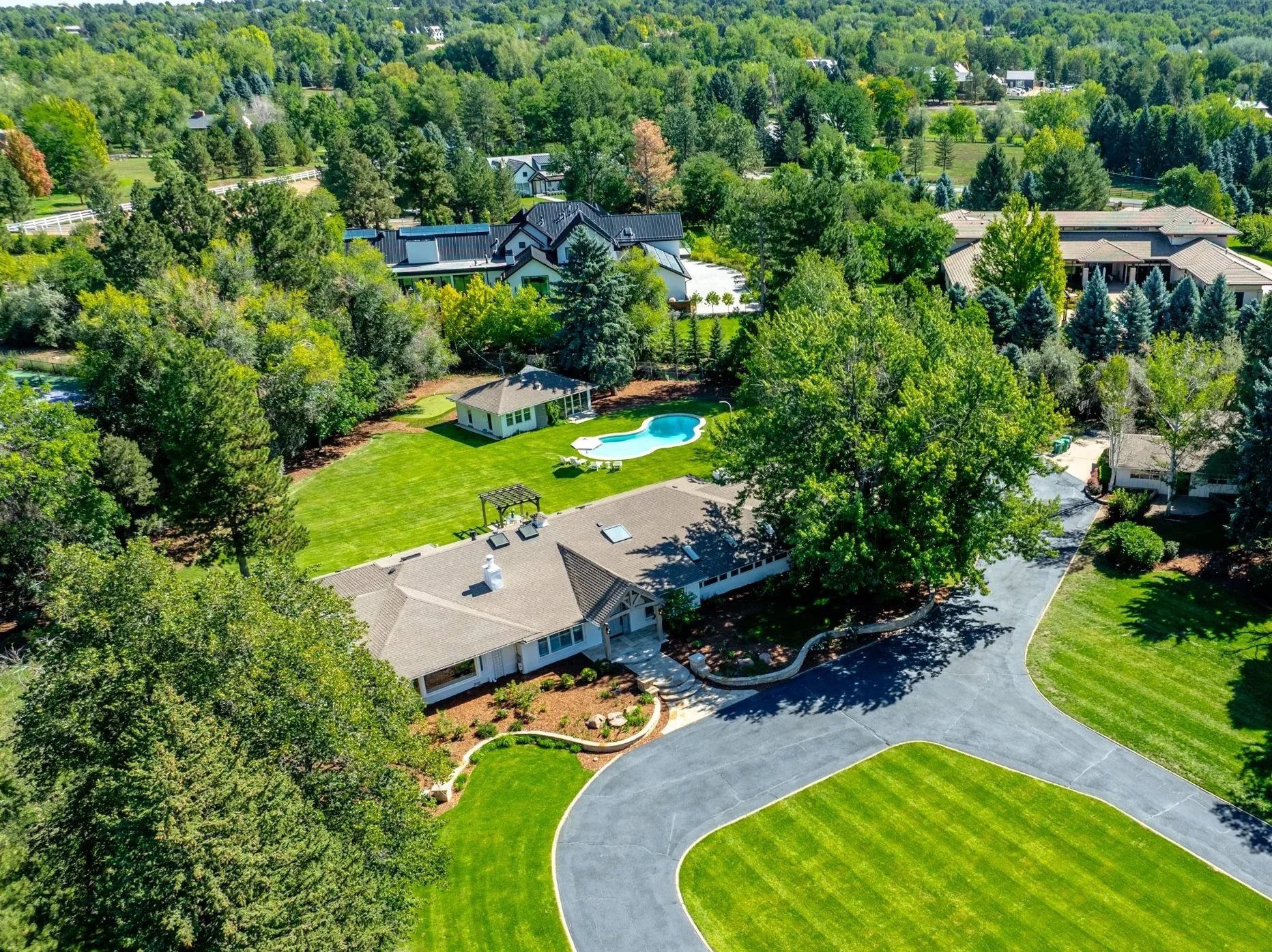 Aerial view of a suburban neighborhood surrounded by green trees. Visible are houses with dark and light-colored roofs, a backyard with a kidney-shaped swimming pool, a lawn, and a driveway. The area is lush with trees and greenery.